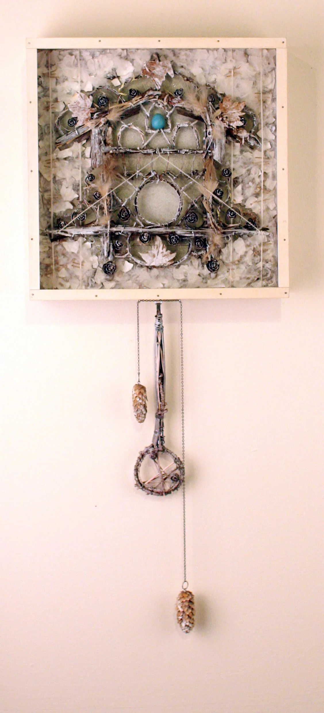 Mixed media wall art piece resembling a snowflake or dreamcatcher, composed of twigs, feathers, pinecones, and natural elements, with a hanging chain and pinecone ornament.