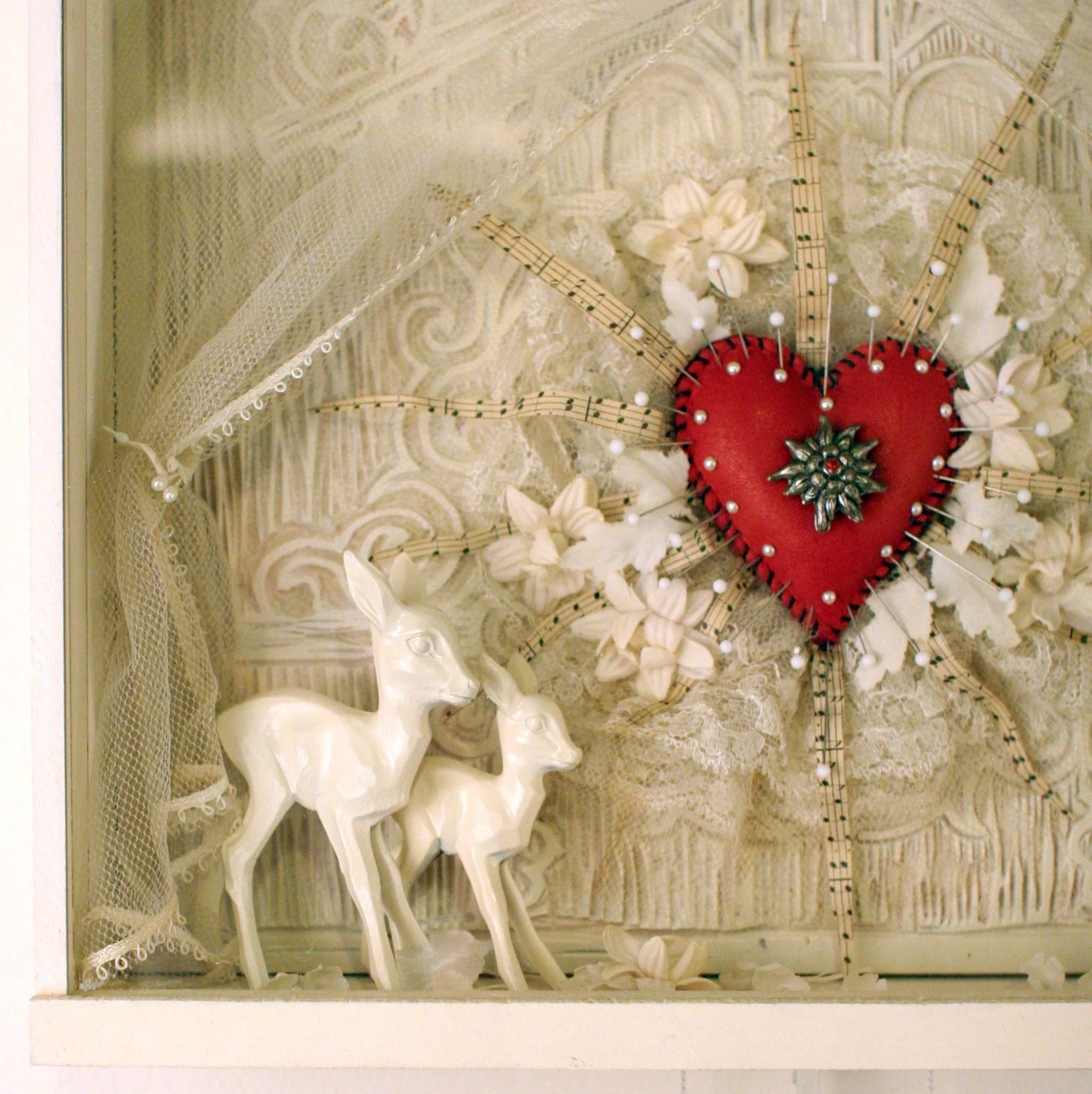 Decorative display featuring a red heart with pins, surrounded by white floral accents, lace, and two white ceramic deer figurines, set against a patterned background.
