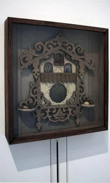 Decorative cuckoo clock with wooden frame and intricate carvings, enclosed in a glass display case on a white wall.