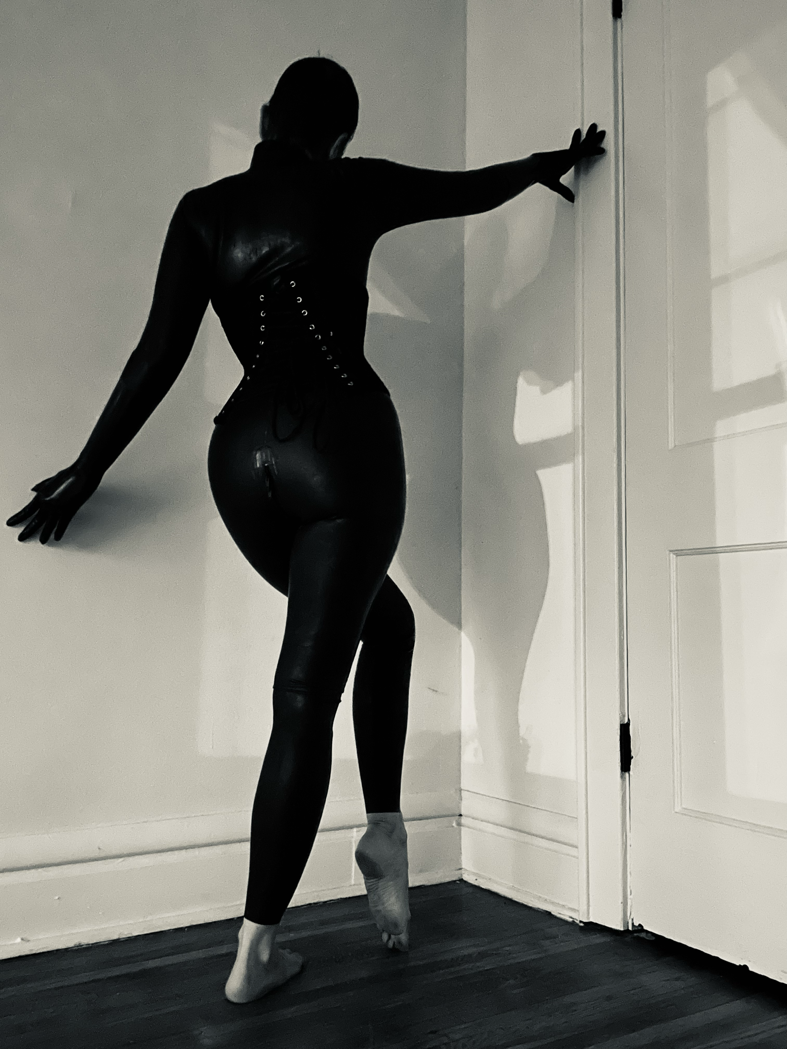 A silhouette of a person in black leather clothing posing with one hand on a door frame and the other on the wall.