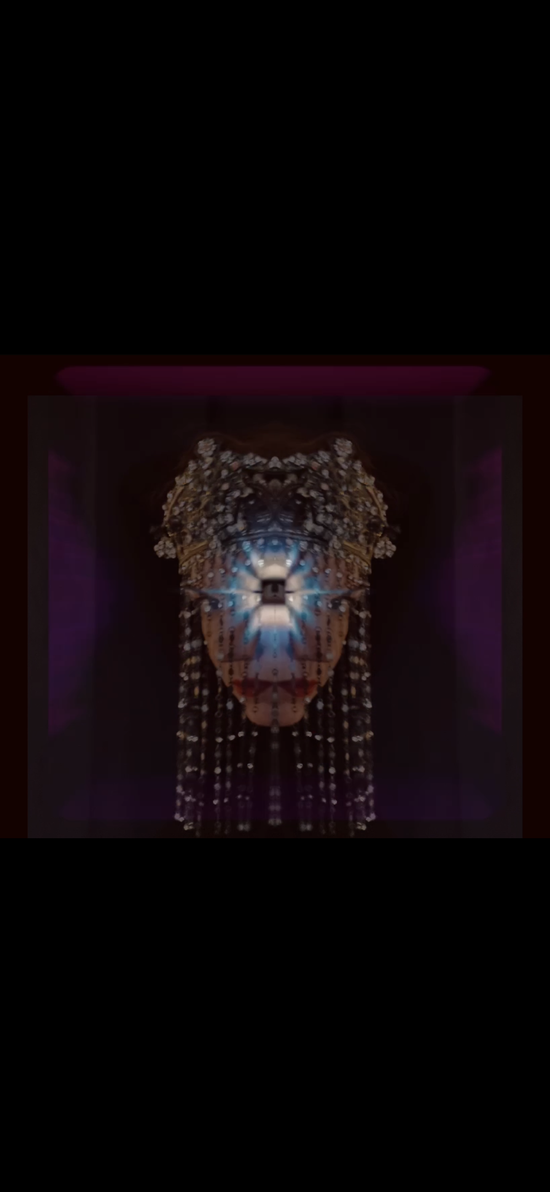 A colorful, symmetrical projection art display featuring intricate patterns and a central glowing light, resembling a face or mask, displayed on a dark wall.