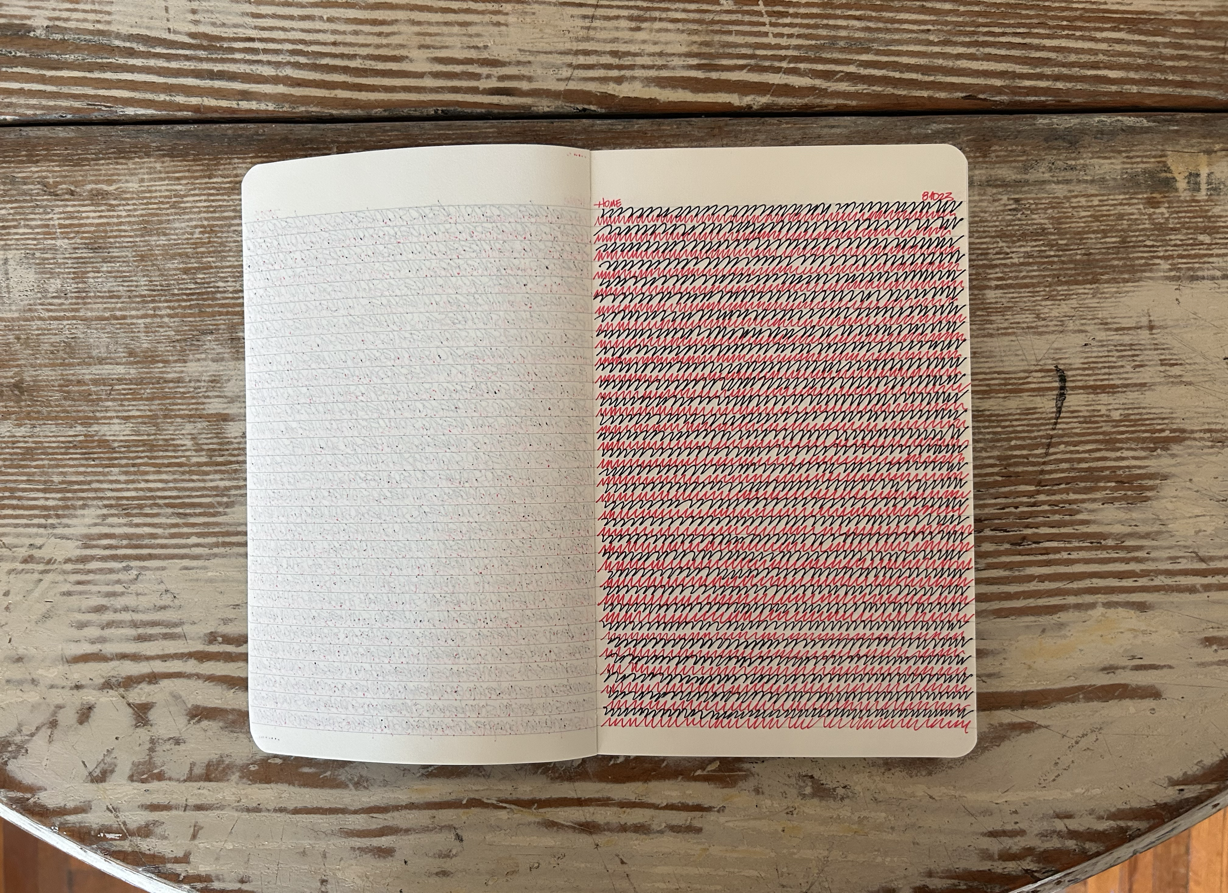 Open notebook on a wooden table with colorful scribbles, red and black lines covering the right page and faint red and blue dots on the left page.