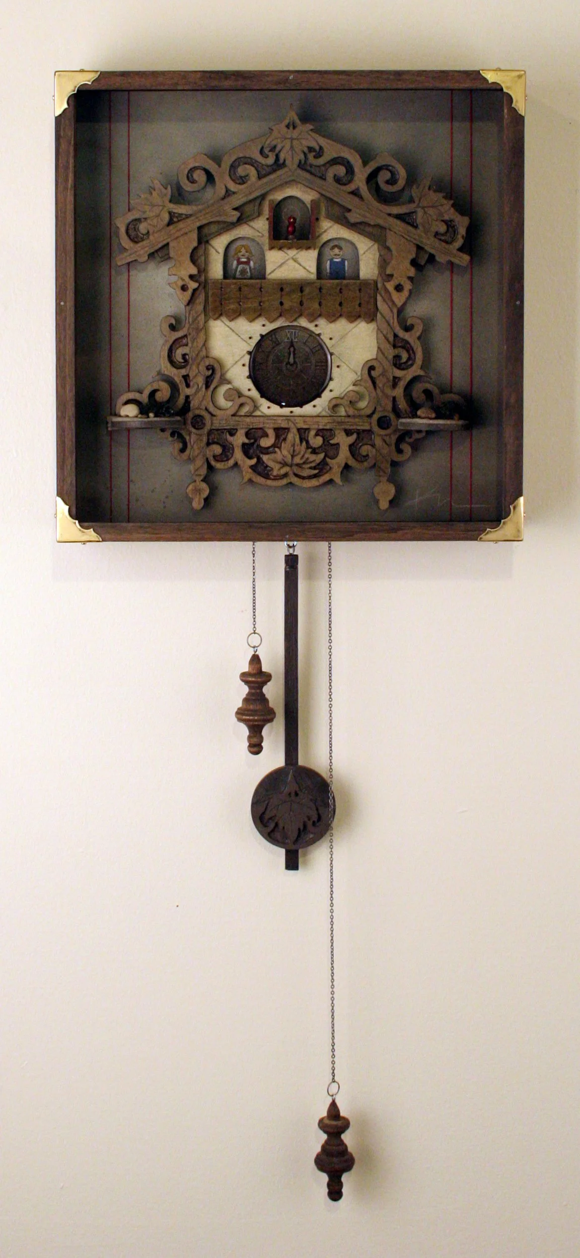 Wooden cuckoo clock with decorative carvings, featuring a small cuckoo bird above the clock face, two figurines inside small doors, and hanging weights below, mounted on a plain wall.