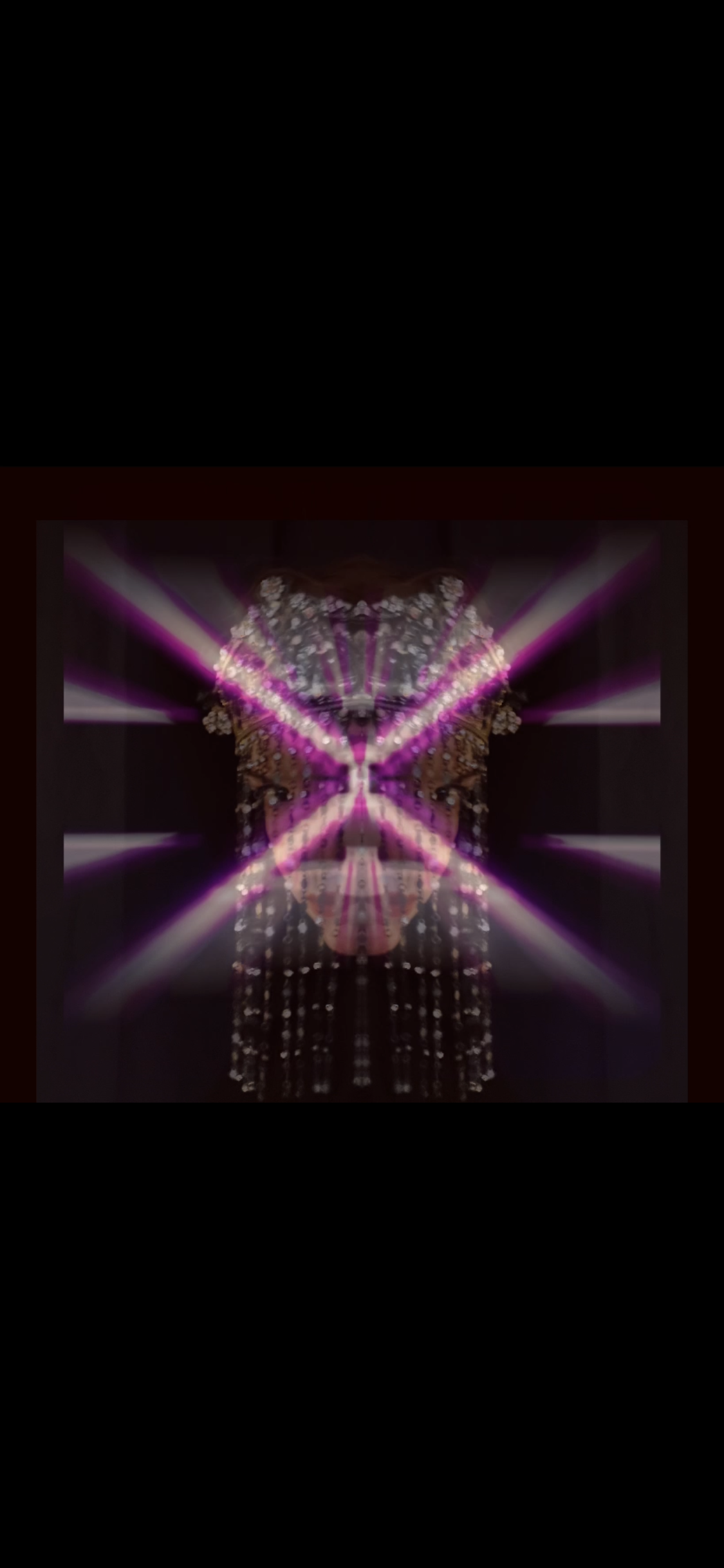 A mirror image of a person with arms raised, standing in front of a light display with pink and white beams forming an 'X' shape, and a backdrop of floral decorations.