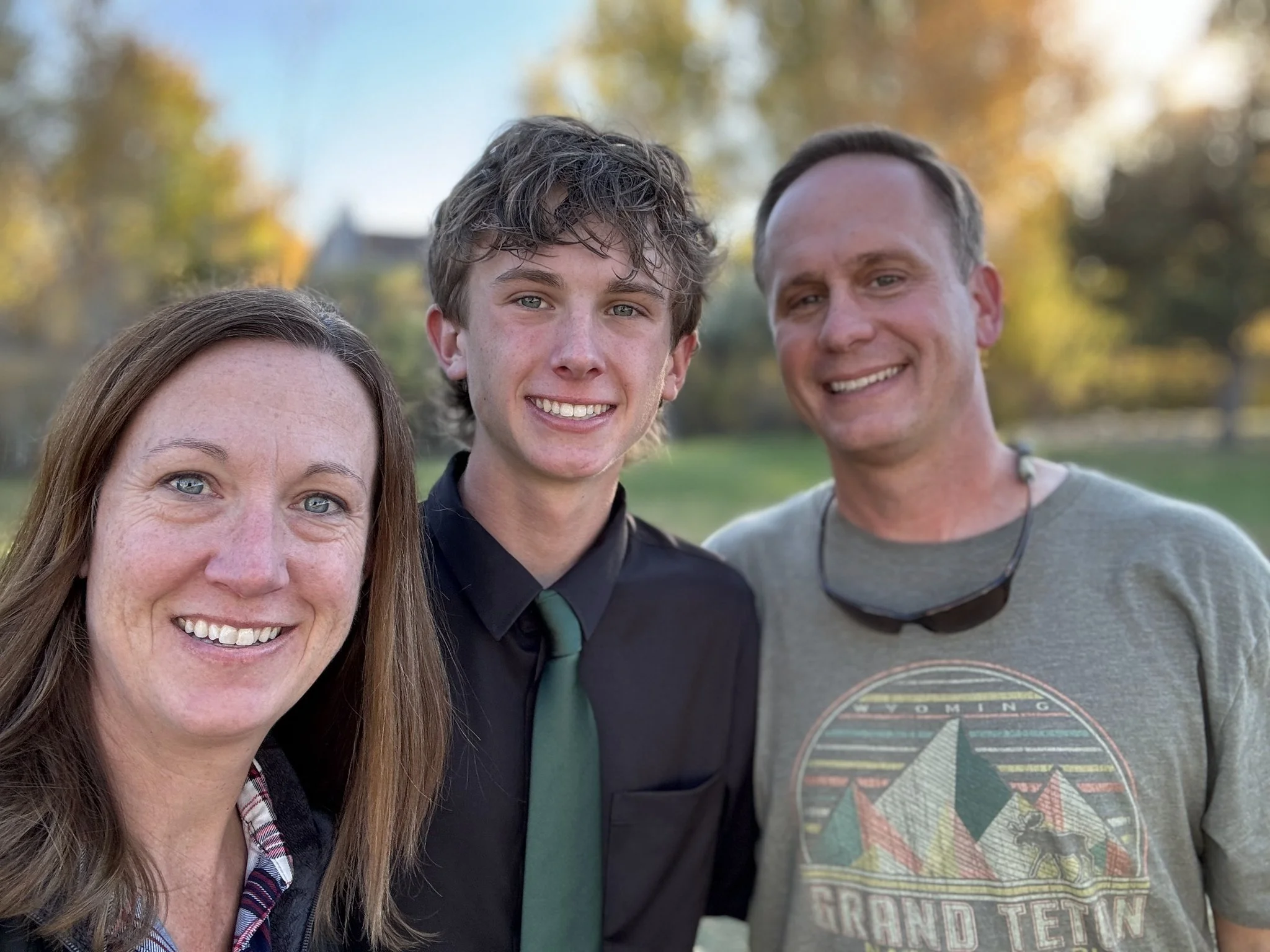 Three smiling people standing outdoors in a park during autumn with trees and fall foliage in the background.