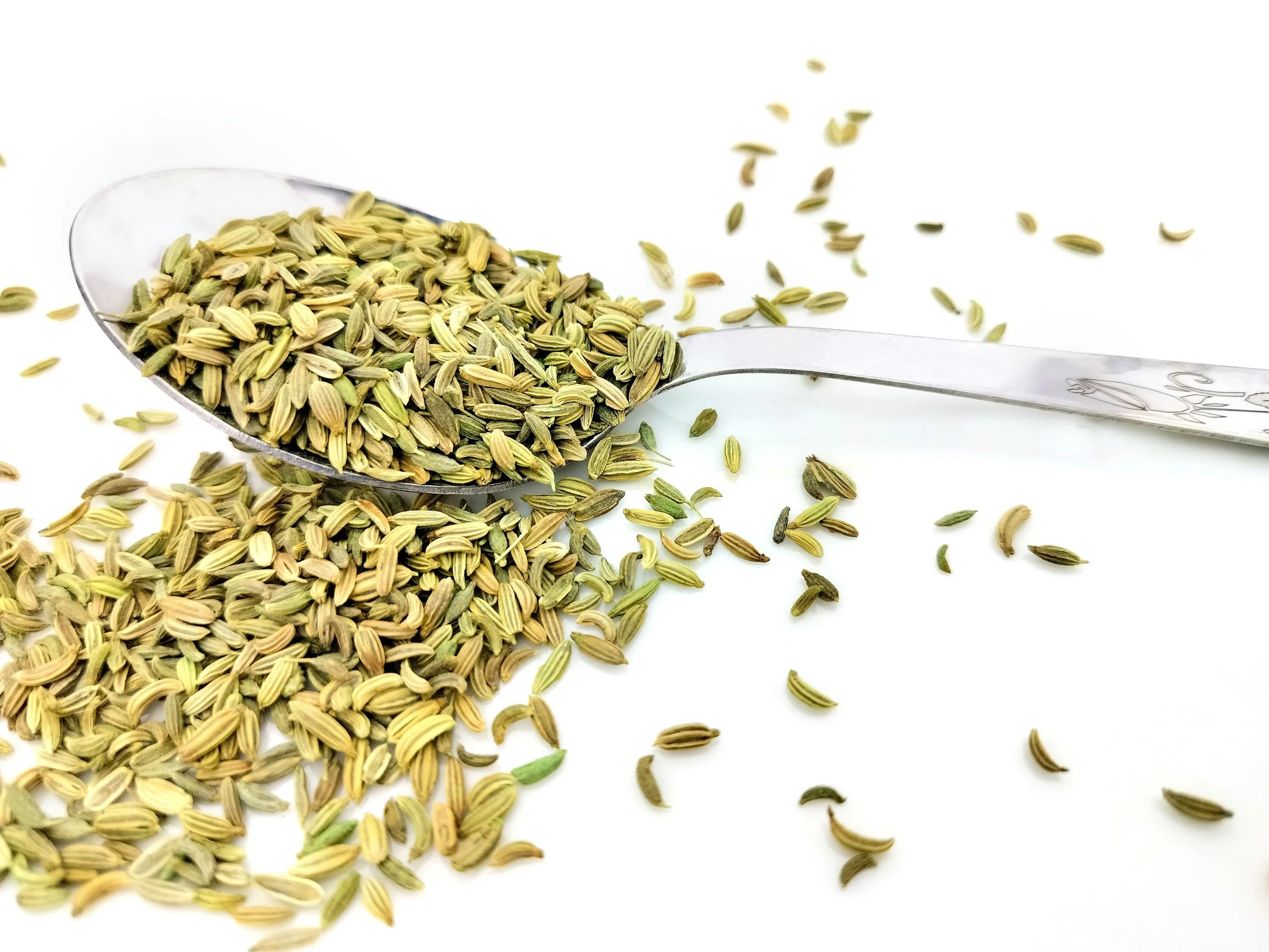A metal spoon filled with dried fennel seeds, with some seeds spilled on a white surface.