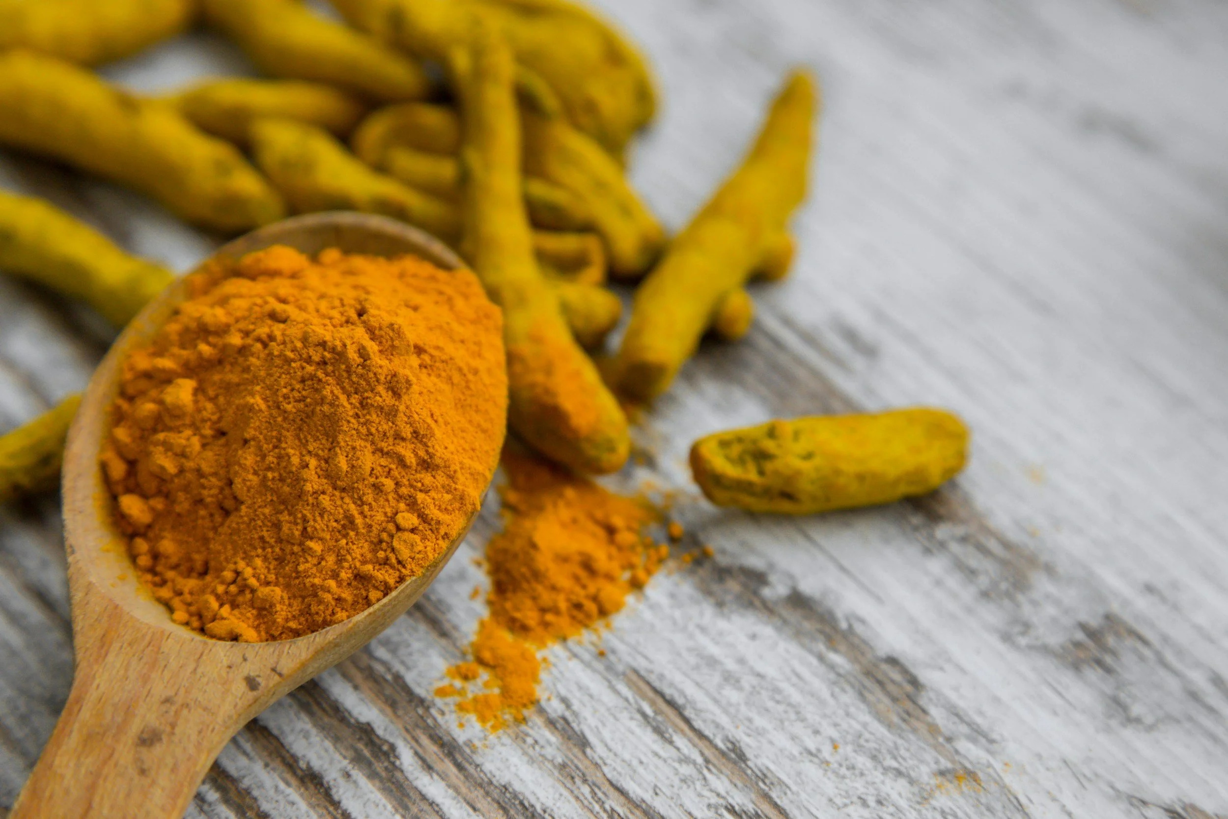Turmeric powder in a wooden spoon surrounded by turmeric roots on a rustic wooden surface.