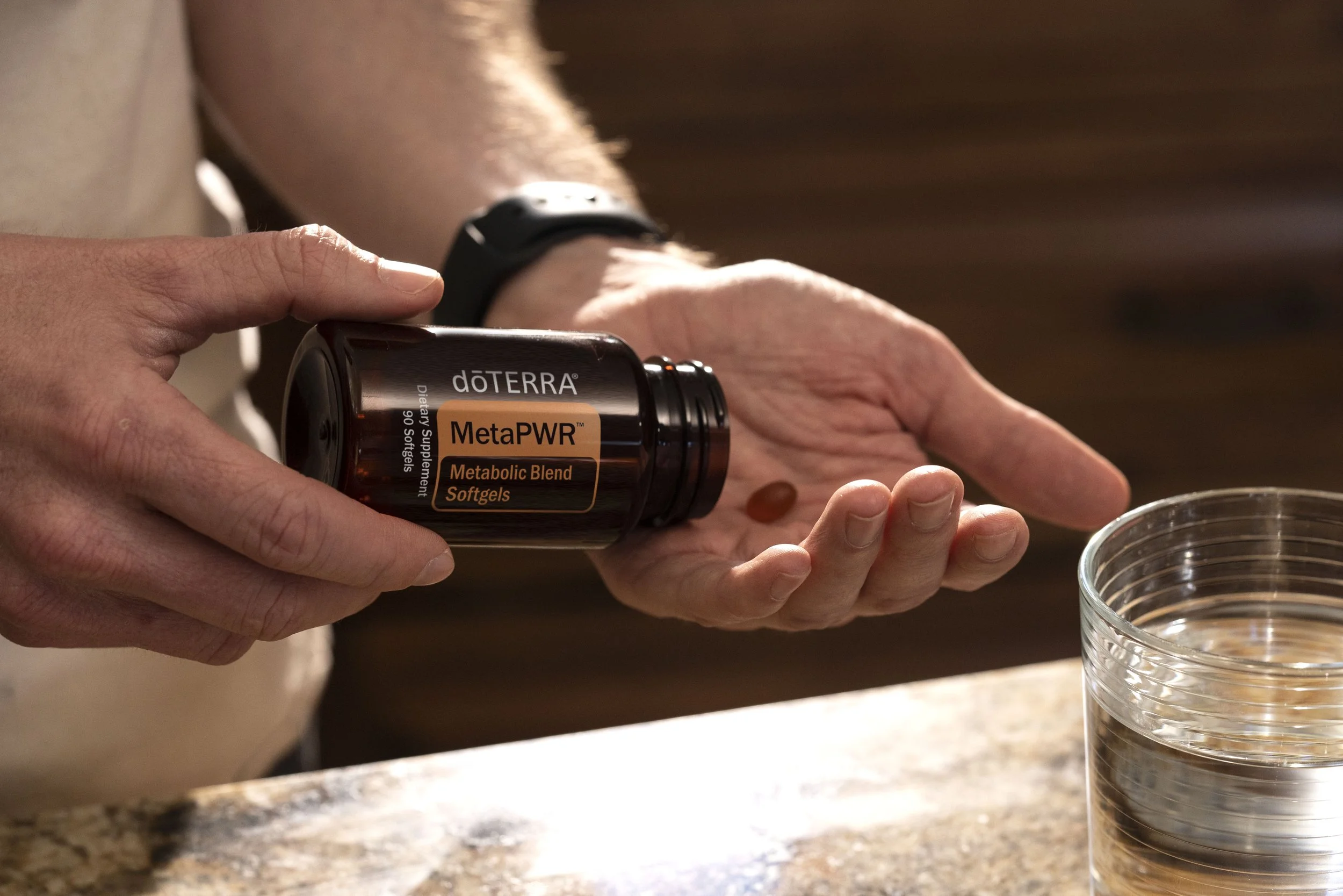 A person with a watch on their wrist holding a bottle of doTERRA MetaPWR Metabolic Blend Softgels, about to pour one softgel into their palm near a glass of water.