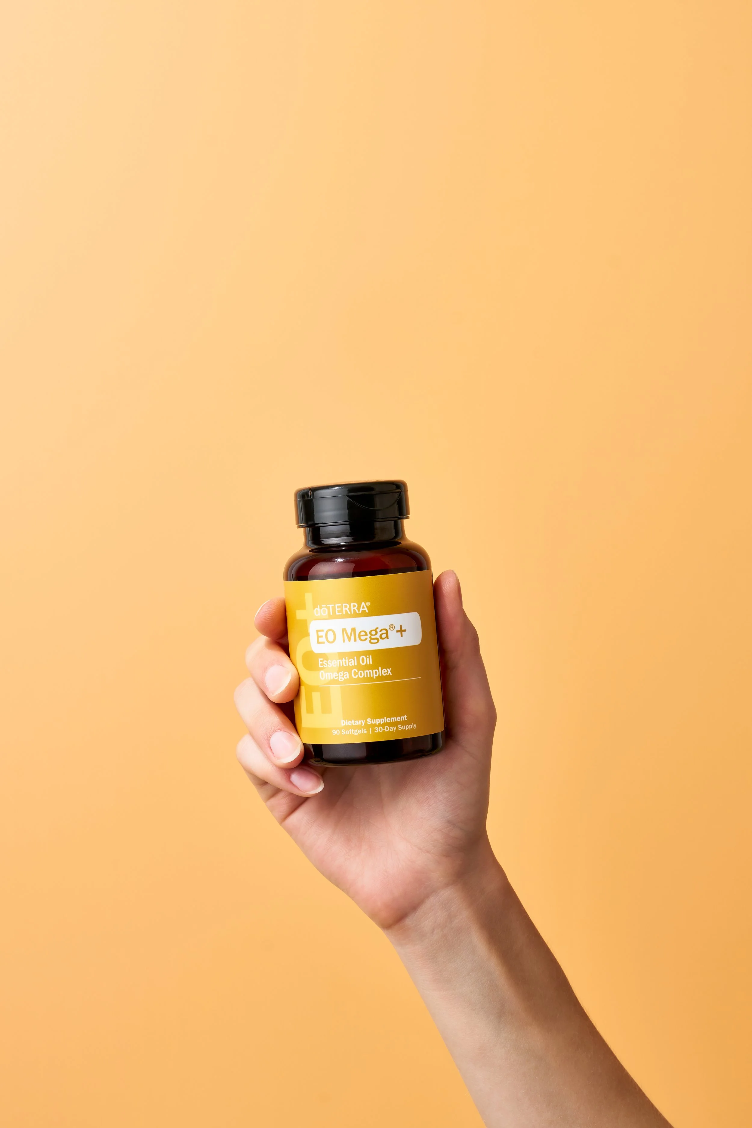 Hand holding a bottle of doTERRA EO Mega+ dietary supplement with a yellow label against a peach-colored background.