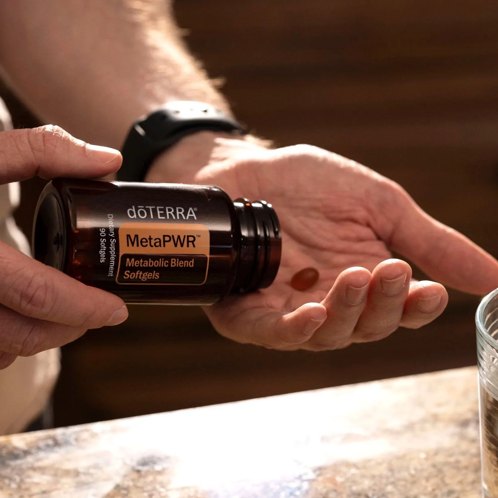 A person with a watch on their wrist is pouring doTERRA MetaPWR dietary supplement softgels from a bottle onto their hand, with a small softgel visible.