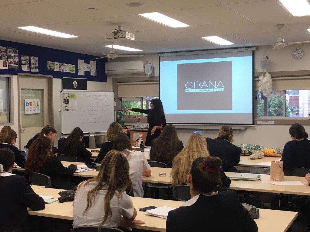 Classroom with students taking notes, teacher pointing at whiteboard, and a screen displaying 'OBANA Fashion Business College'