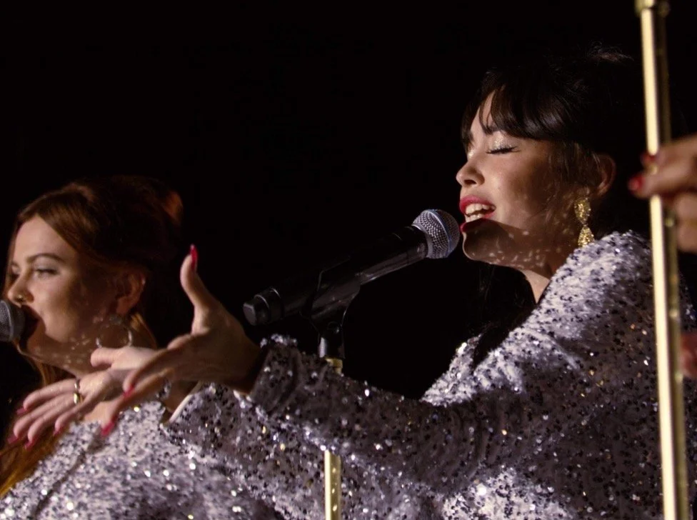 Two women singing into microphones at a performance, one in a sparkly silver dress with eyes closed, the other in the background.
