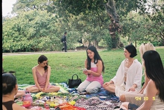 Group of women sitting cross-legged on a picnic blanket during a picnic in a park with trees and grass.