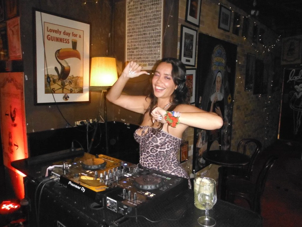 A woman with long dark hair wearing a leopard print dress and a red floral bracelet is smiling and dancing behind DJ equipment in a dimly lit bar or nightclub.