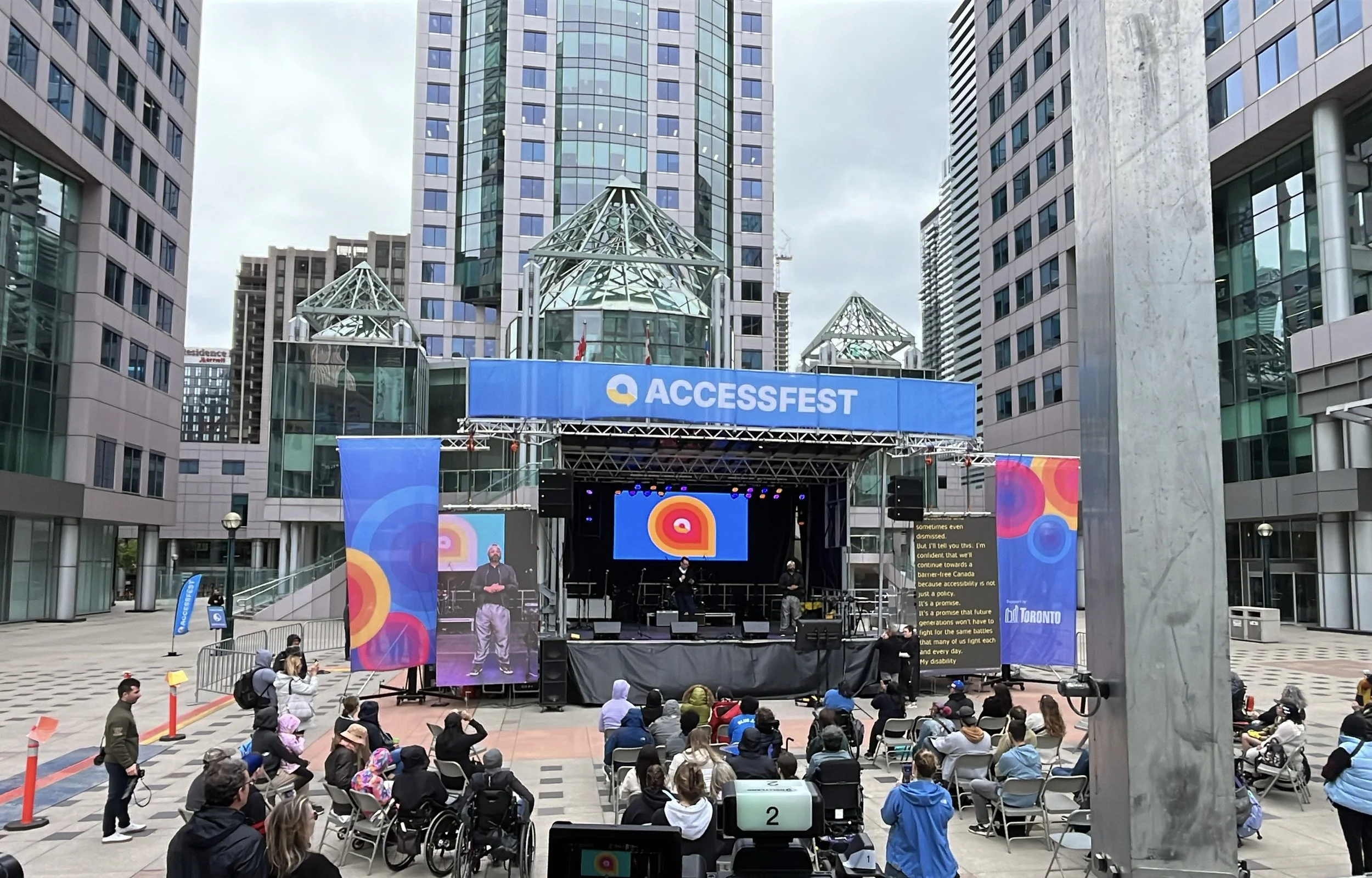 An outdoor stage with the banner 'ACCESSFEST' in the center, set up in an urban plaza surrounded by tall modern buildings. The stage features a large screen displaying a colorful logo, with several speakers and stage lights. An audience, some in whee