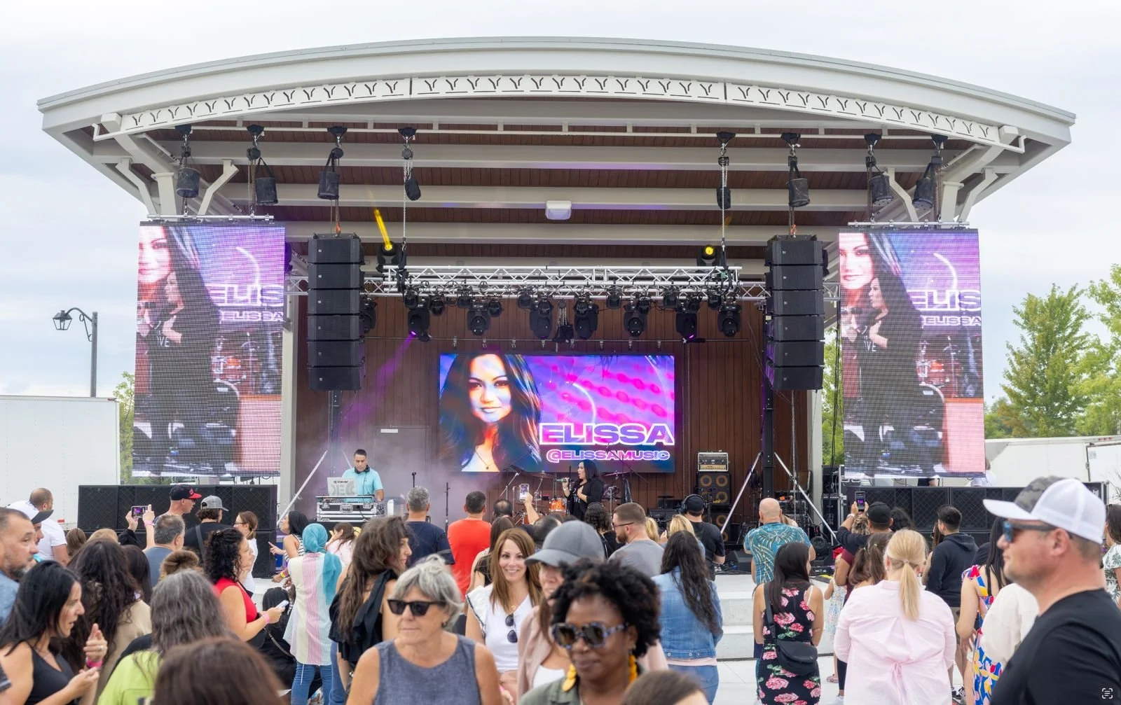 An outdoor music concert with a large stage, featuring a digital screen displaying a woman’s face and the name 'Elissa' with the social media handle '@Elissamusic'. There are two tall vertical screens on each side of the stage, multiple speakers, and