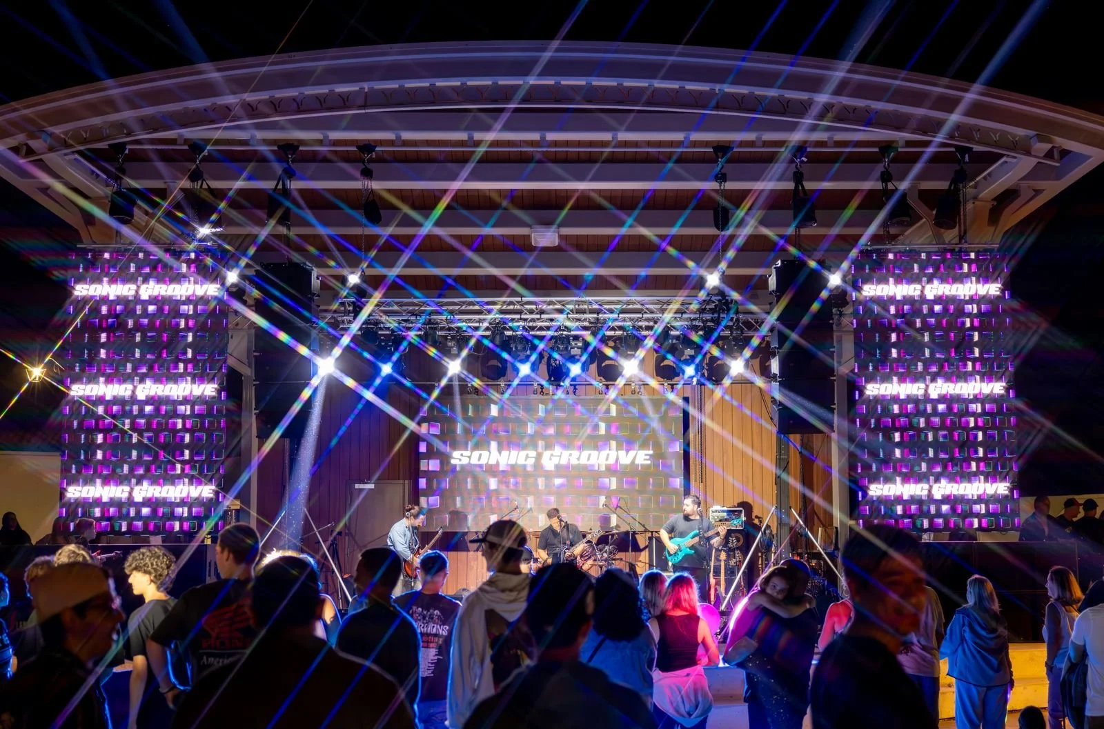 A live music concert with a band performing on stage under colorful lighting, with a crowd of people watching and dancing in front.