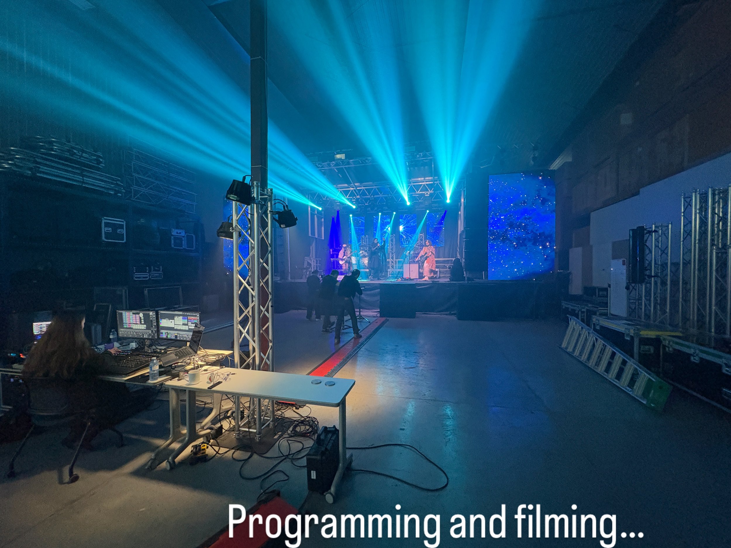 An indoor concert venue with a stage lit by blue and purple lights, musicians performing, a person working on a sound mixing console, and a few people walking around.