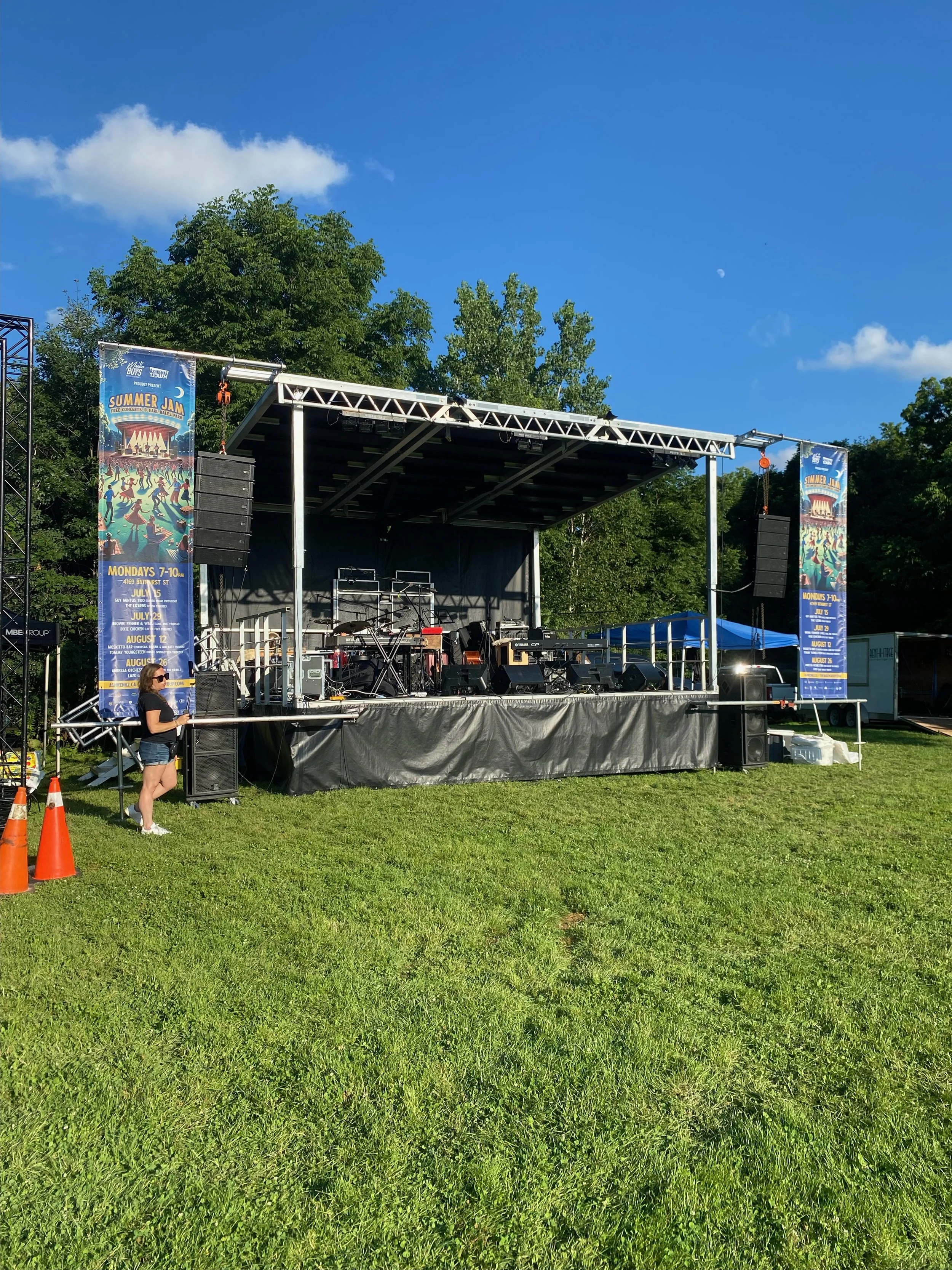 An outdoor stage set up for a summer music event with banners on each side, musical instruments, and equipment, with a woman standing nearby, set against a background of trees and a bright blue sky.