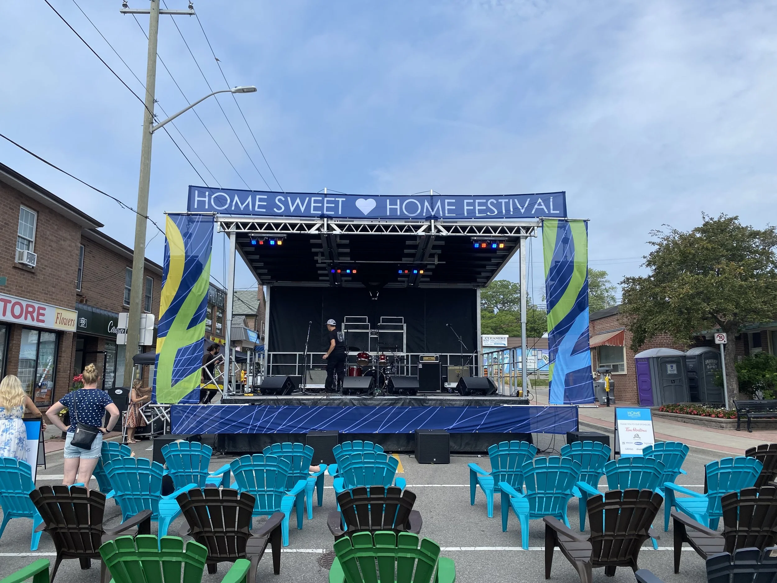 An outdoor stage set up for the Home Sweet Home Festival, with chairs arranged in front, some people setting up, and a few performers on stage, under a blue sky with a few clouds.