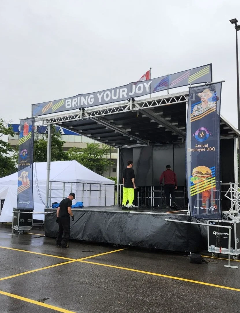 An outdoor event stage with a sign that reads 'BRING YOUR JOY'. There are two vertical banners advertising an annual employee BBQ. The stage has three people on it and is set in a parking lot on a rainy day, with tents and trees in the background.