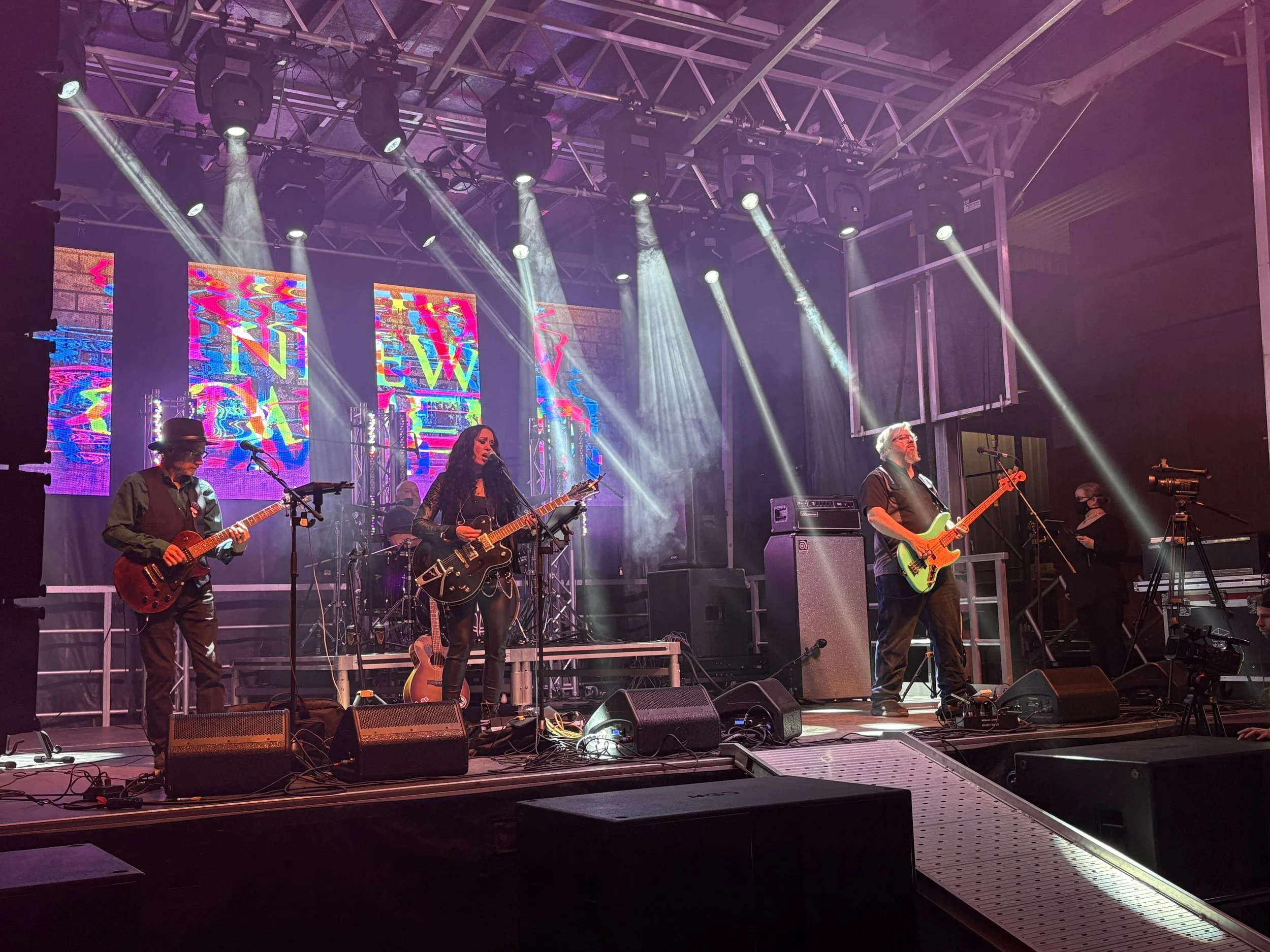A band performing on stage with colorful digital screens in the background, featuring four musicians playing guitars and a drummer, with stage lighting and audio equipment.