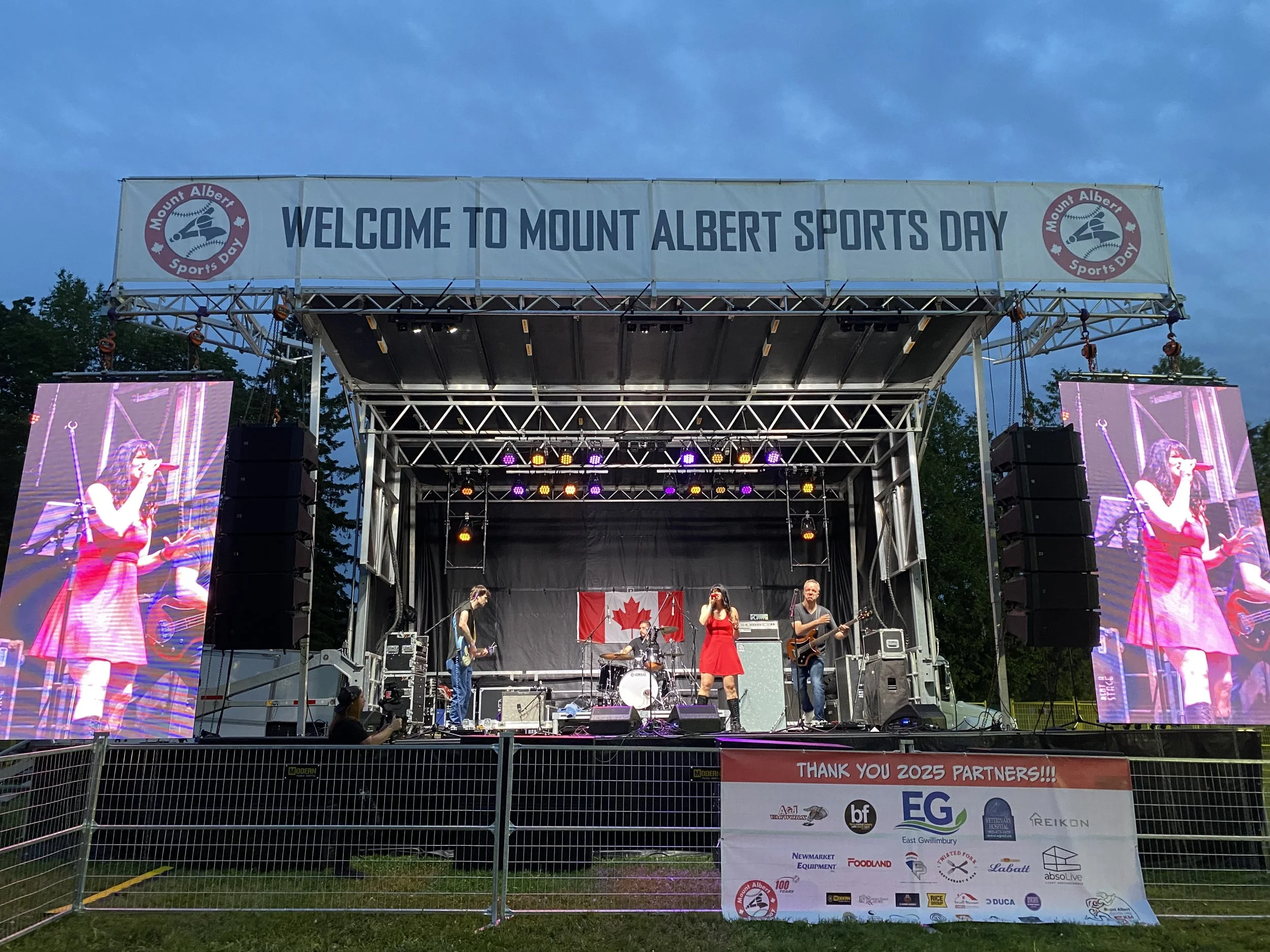 Stage at Mount Albert Sports Day with a band performing, Canadian flag in the background, and two large screens displaying the performers.