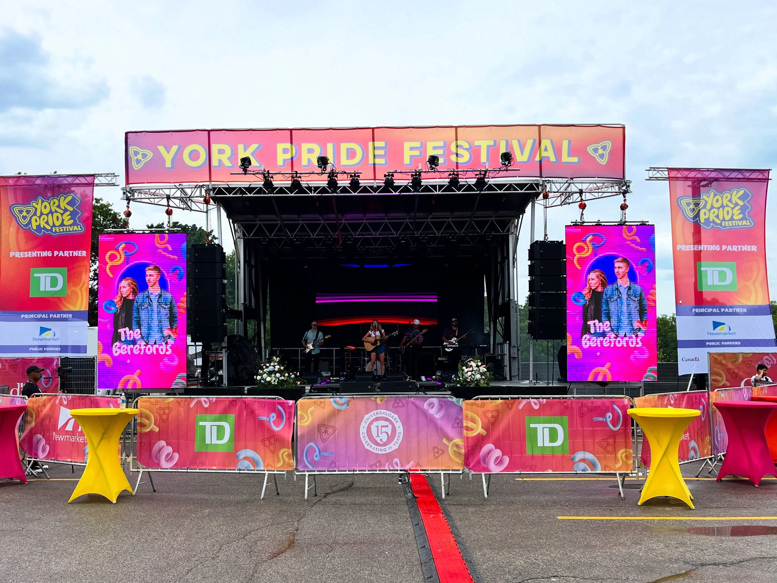 Stage at the York Pride Festival with a band performing, decorated with colorful banners and digital screens, in an outdoor setting under a cloudy sky.