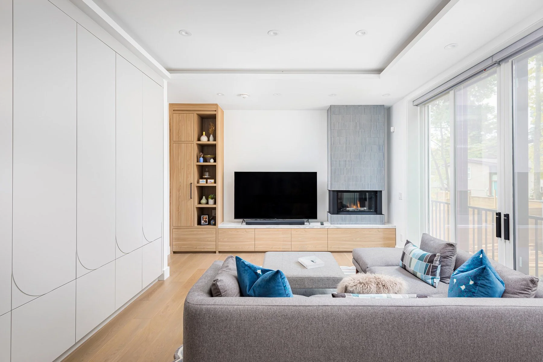 Modern living room with a gray couch, colorful pillows, a flat-screen TV, a wood cabinet, and large glass sliding doors with a balcony view.