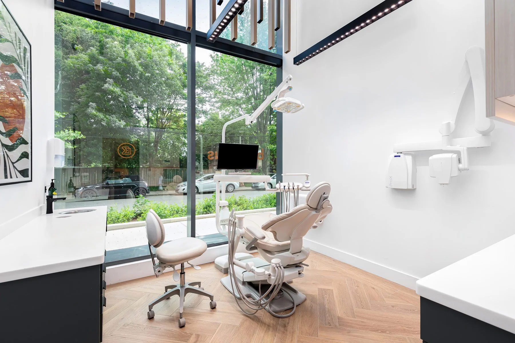 Modern dentist's office treatment room with dental chair, tools, monitor, and large window showing green trees outside.