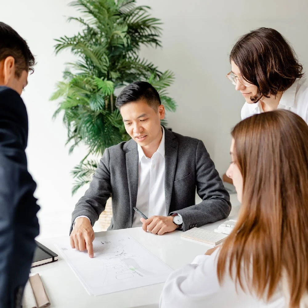 Group of four professionals in a business meeting discussing plans or designs at a white table, with a potted plant in the background.