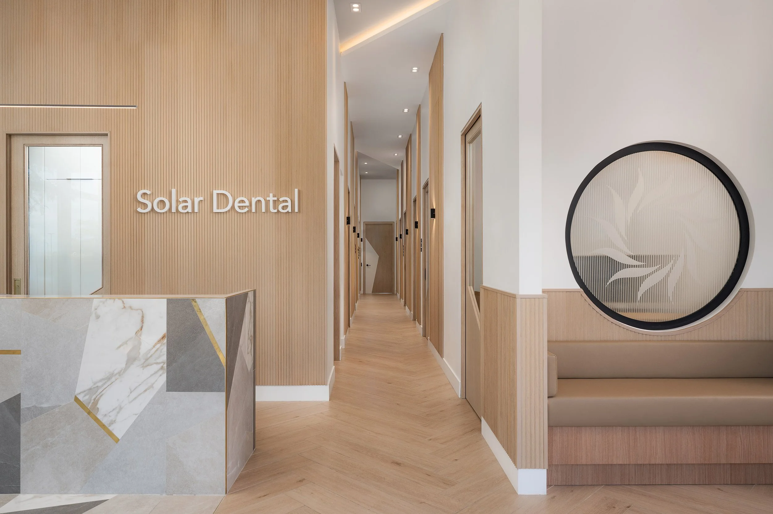 Modern dental clinic interior with wooden accents, a reception desk with marble pattern, hallway with room doors, and a decorative circular wall art.