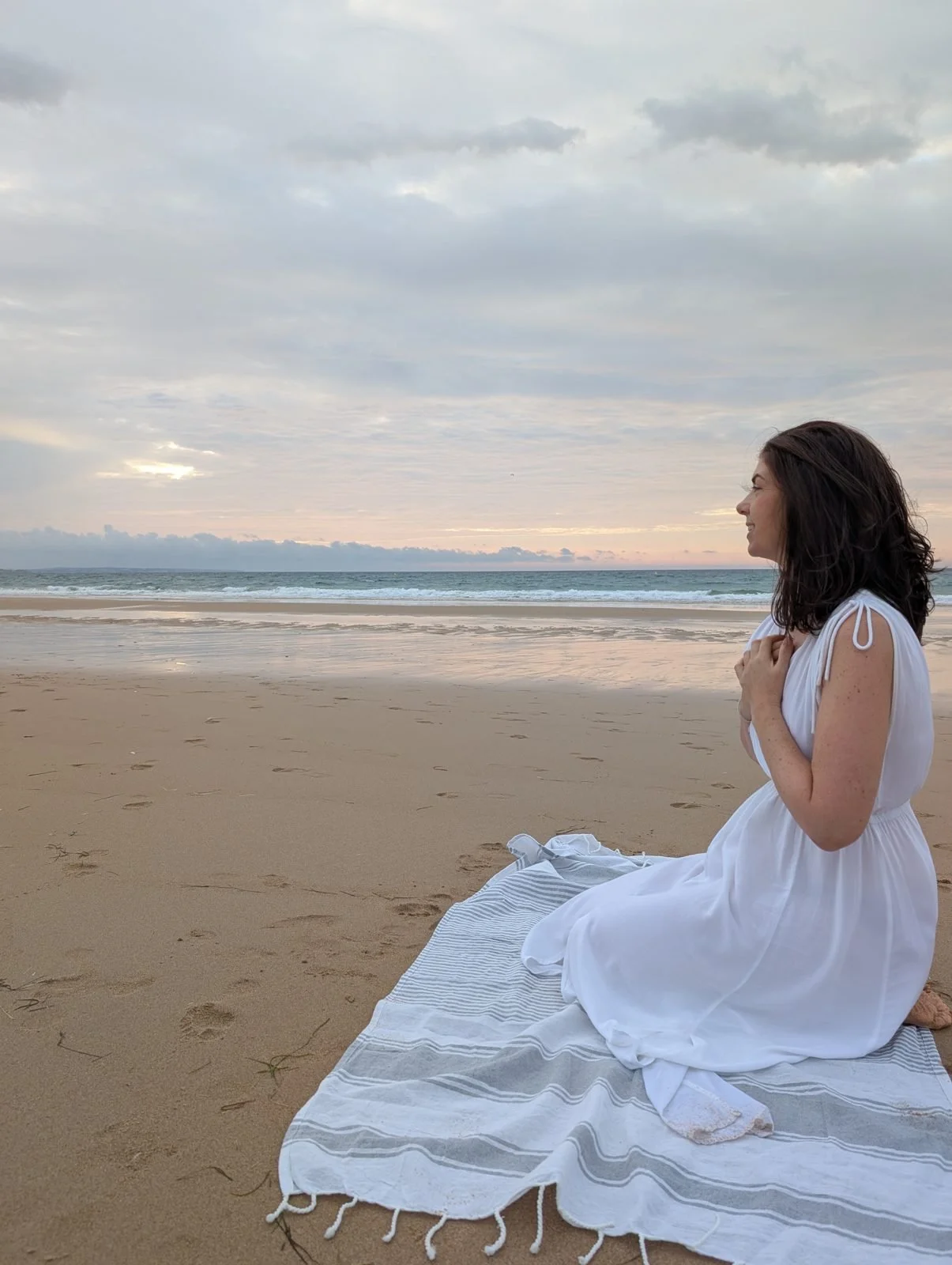 Katie Boysen sitting on a beach in the Algarve at sunrise with her hands on her heart