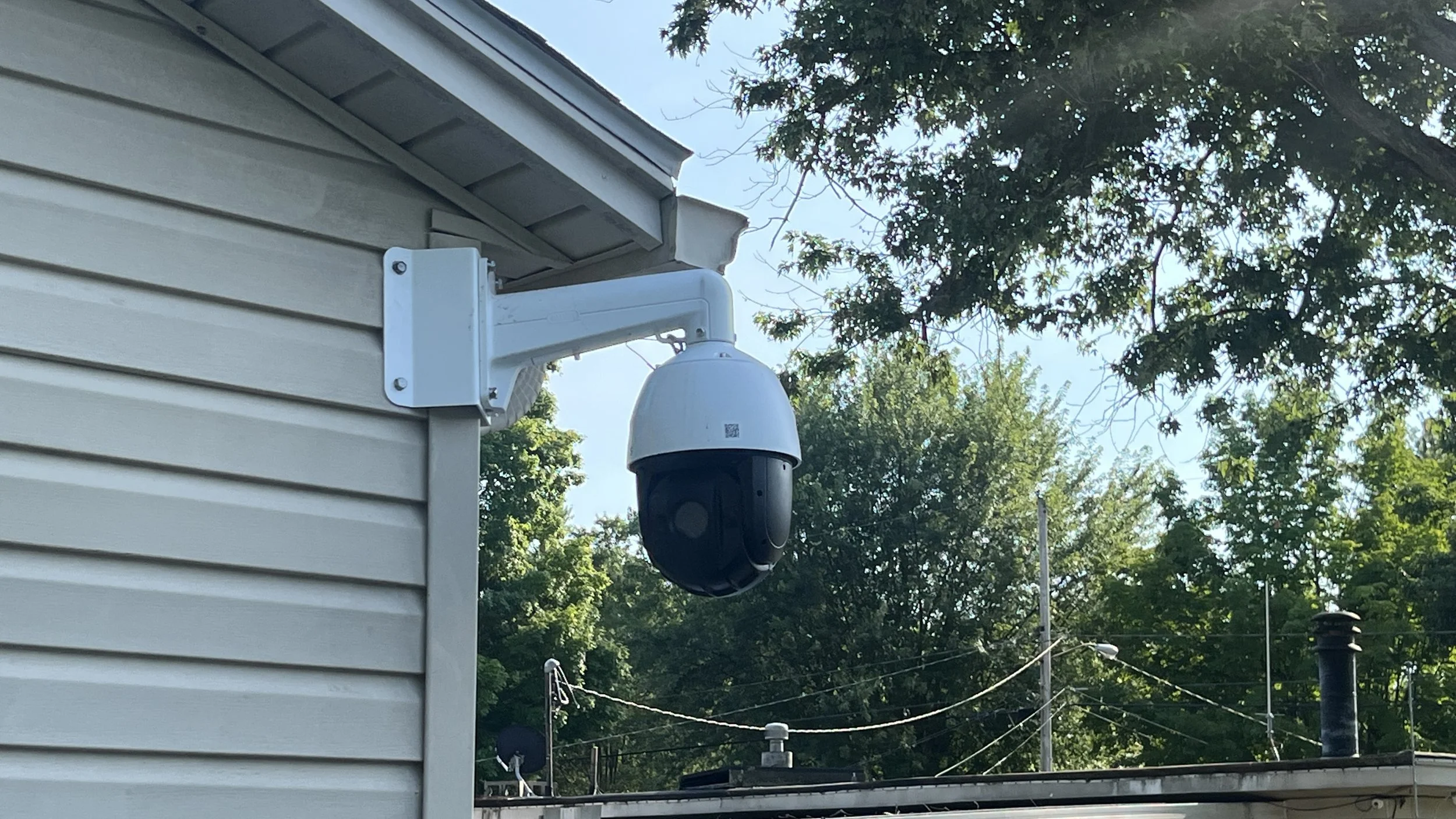 Security camera mounted on the side of a house, with trees and power lines in the background.