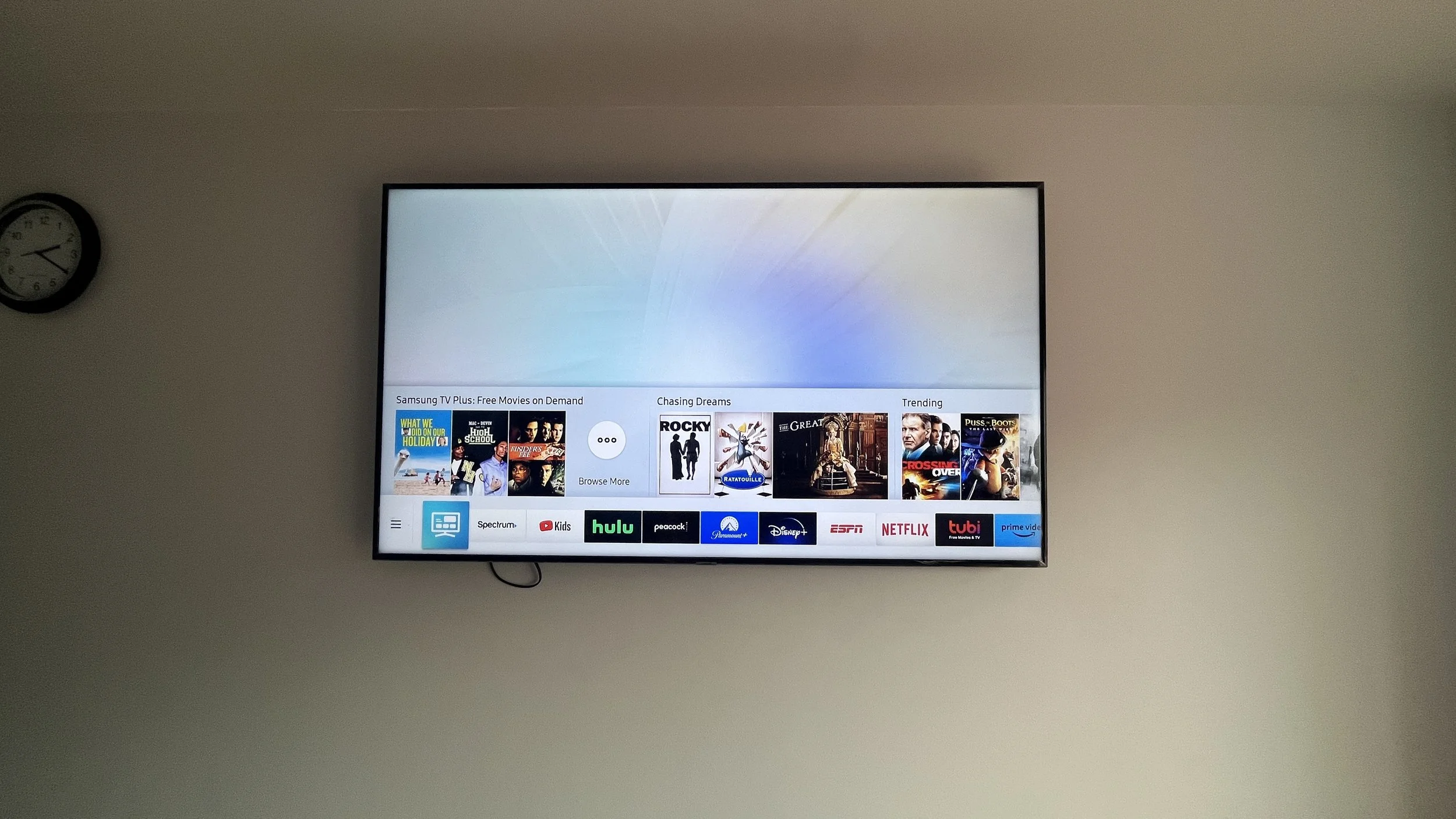 Television screen displaying a menu of streaming services and movies, mounted on a white wall beside a clock showing 7:07.