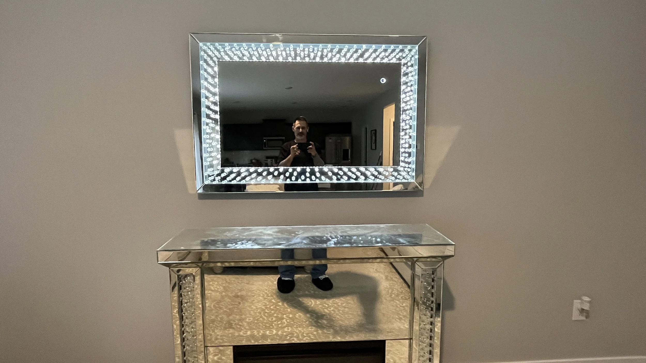 A man taking a photo of himself in a decorative mirror with LED lights. The mirror is mounted on a beige wall above a mirrored console table.