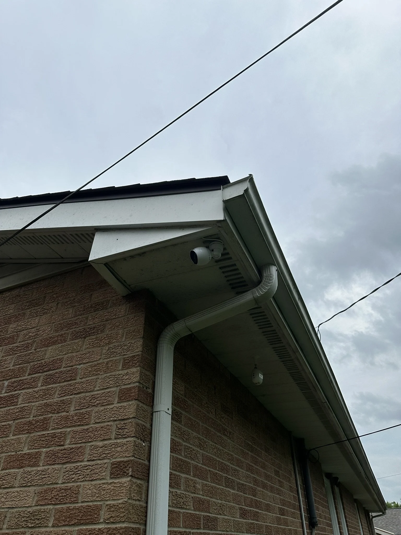 Close-up of the corner of a brick house showing a white security camera below the roof eave, a white gutter, and drain pipe against an overcast sky.