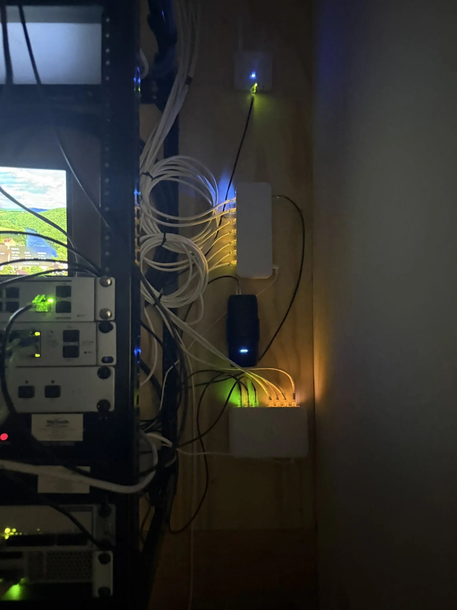 Rack of networking equipment with numerous cables connected, power strips, and devices emitting green, blue, and yellow lights on a wooden floor.