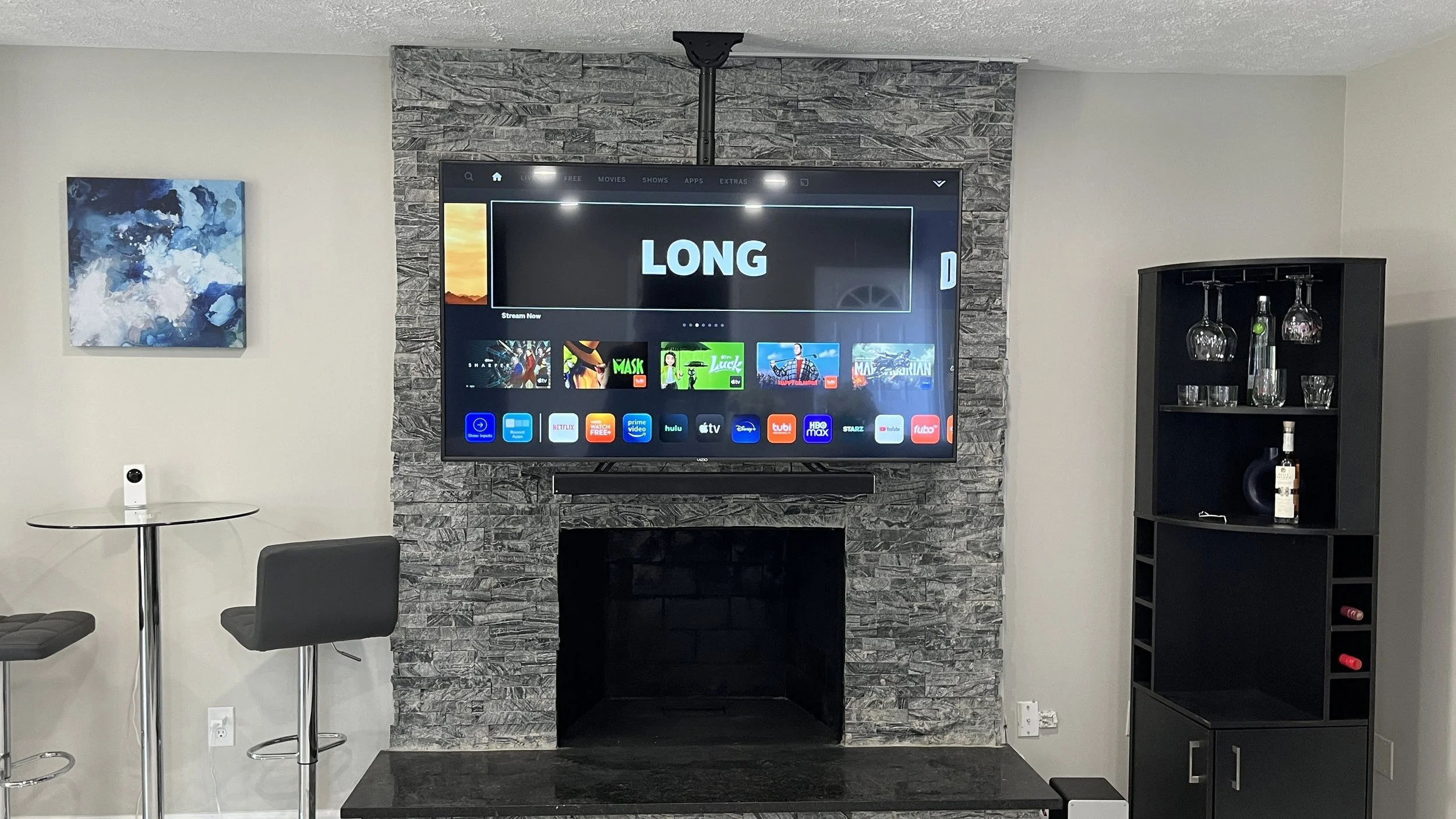 Living room with a large flat-screen TV displaying a streaming app, mounted on a textured stone wall above a fireplace. To the left, a small round table with a security camera and a black chair. To the right, a black cabinet with glassware and bottles, including a prominent bottle of alcohol.