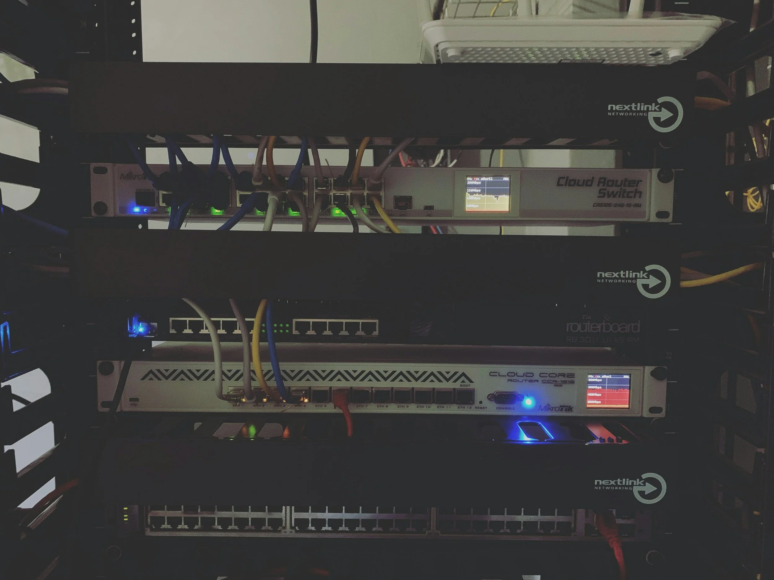 A rack of networking equipment including routers, switches, and fiber optic modules with illuminated indicator lights and a small display screen, all arranged in metal enclosures.