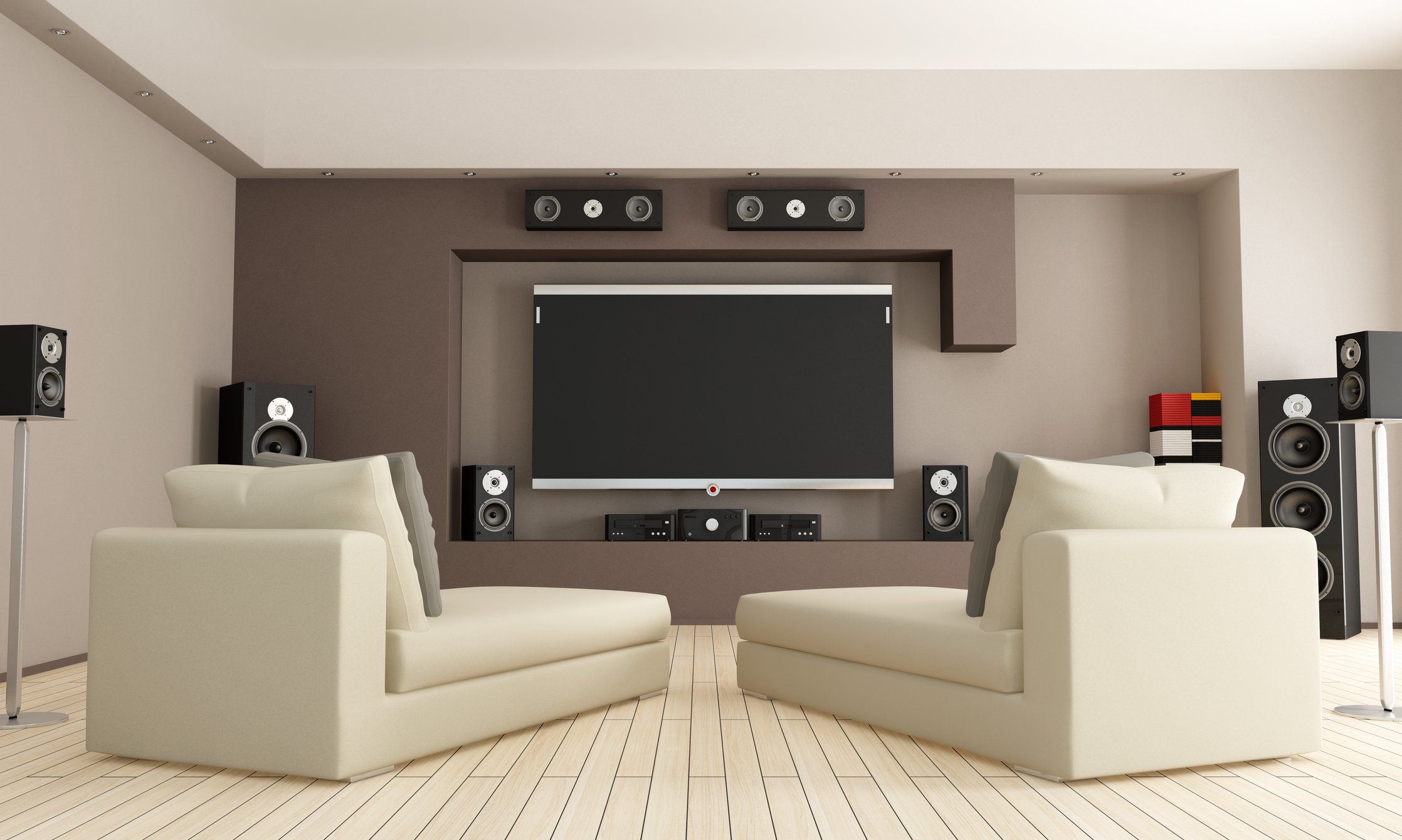 A modern home theater room with white leather sofas, a large flat-screen TV, multiple black surround sound speakers on the walls and ceiling, light wood flooring, and a small white table.