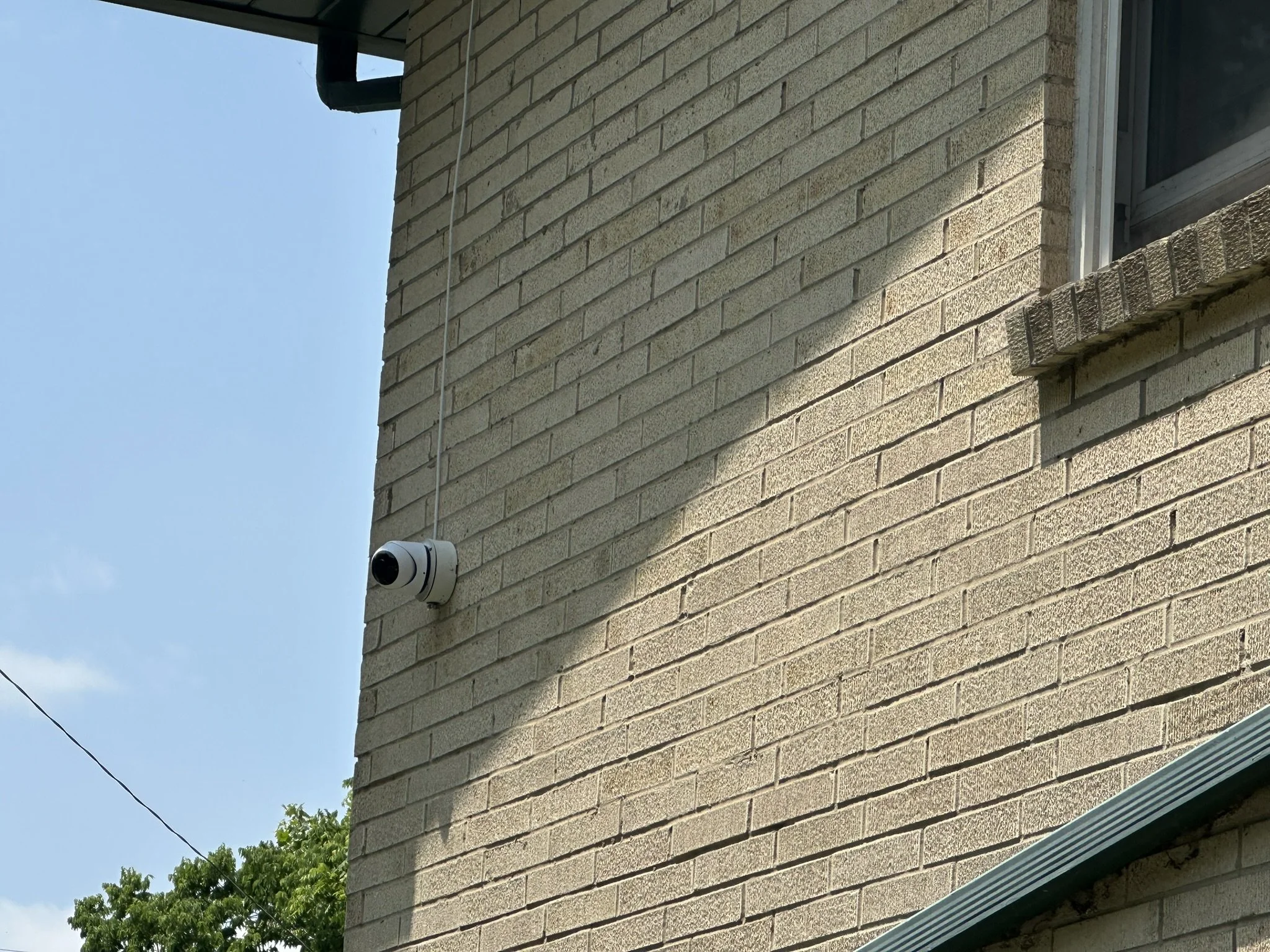 Security camera mounted on the exterior wall of a house with a brick facade, under a partially visible roof and a clear blue sky.