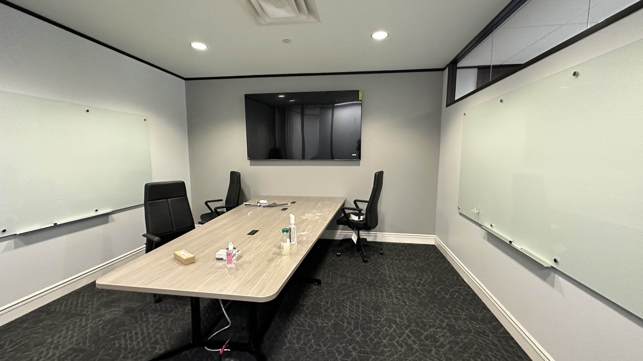 Empty conference room with a rectangular table, three black chairs, whiteboards on the walls, a large wall-mounted TV, and various items on the table.