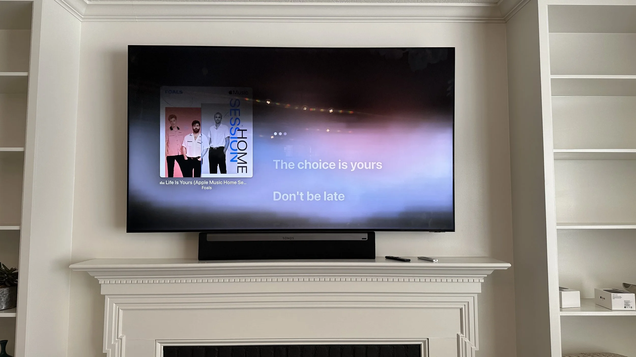Television screen displaying the song 'Life Is Yours' by Foals with album cover art showing three band members, and lyrics 'The choice is yours' and 'Don't be late'.
