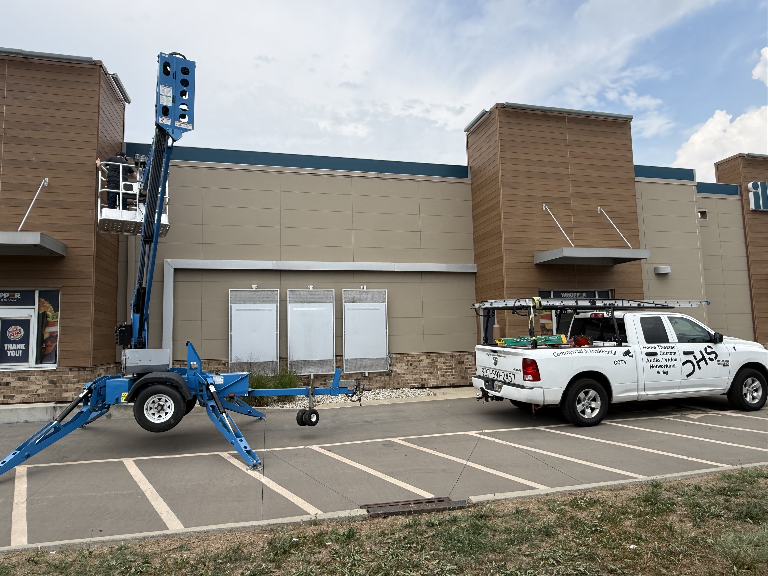 Construction worker on a blue lift working on the exterior of a commercial building with beige and brown siding under a partly cloudy sky.