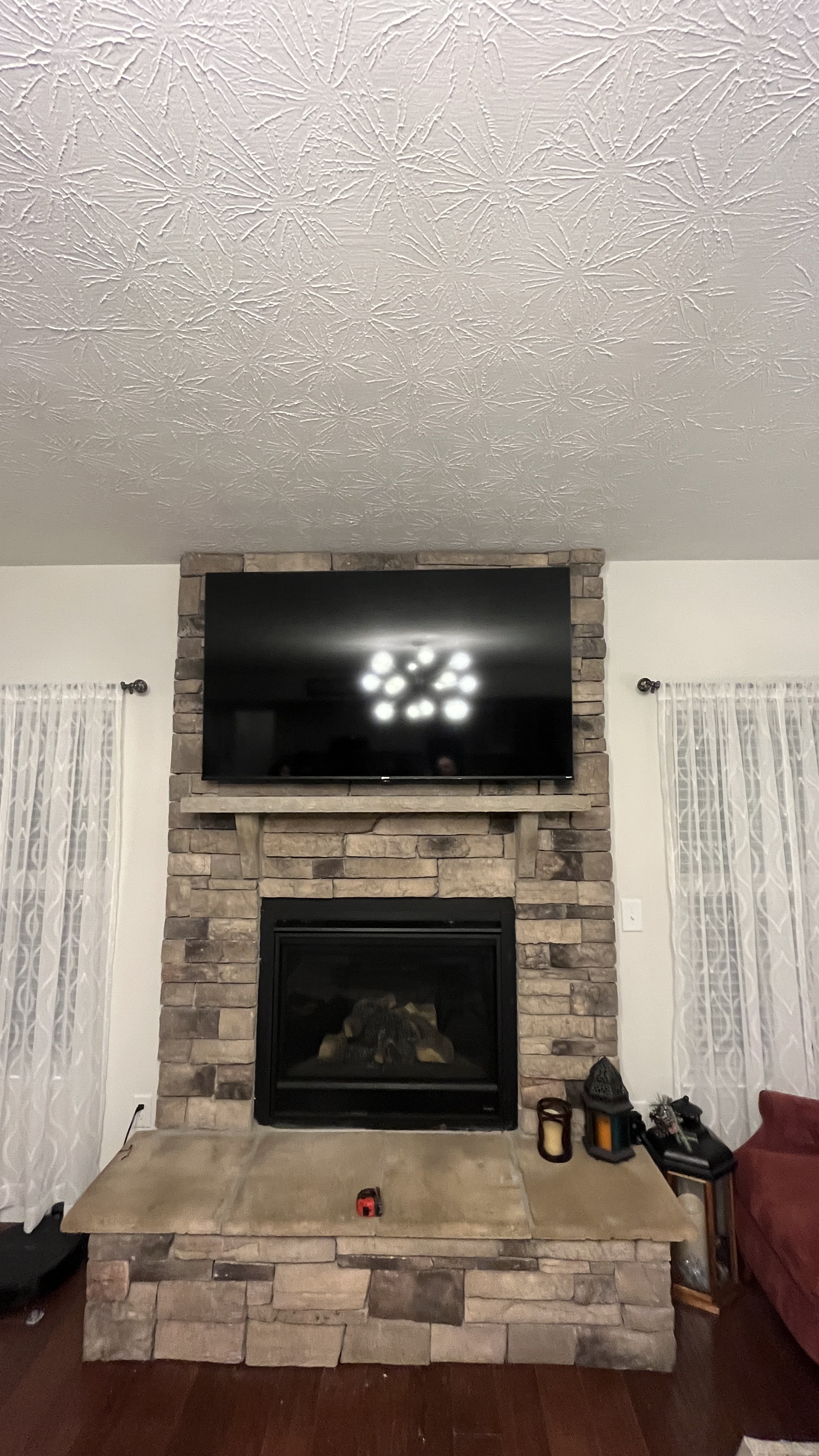 Living room fireplace with a stone surround, a mounted flat-screen TV above, and a beige hearth with various items on it, flanked by patterned white curtains.