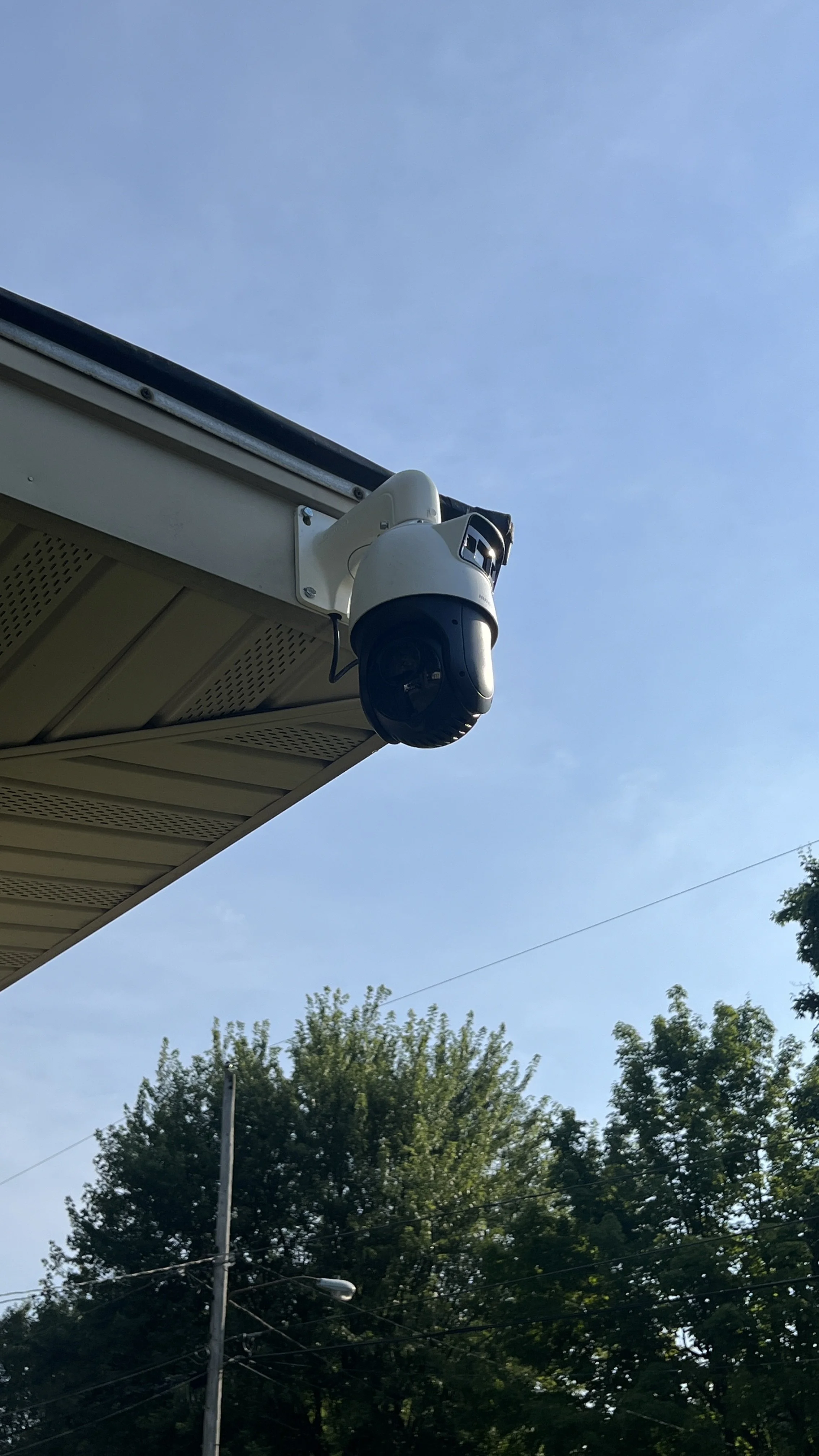Security camera mounted under the eaves of a building, with trees and power lines in the background and a clear blue sky.