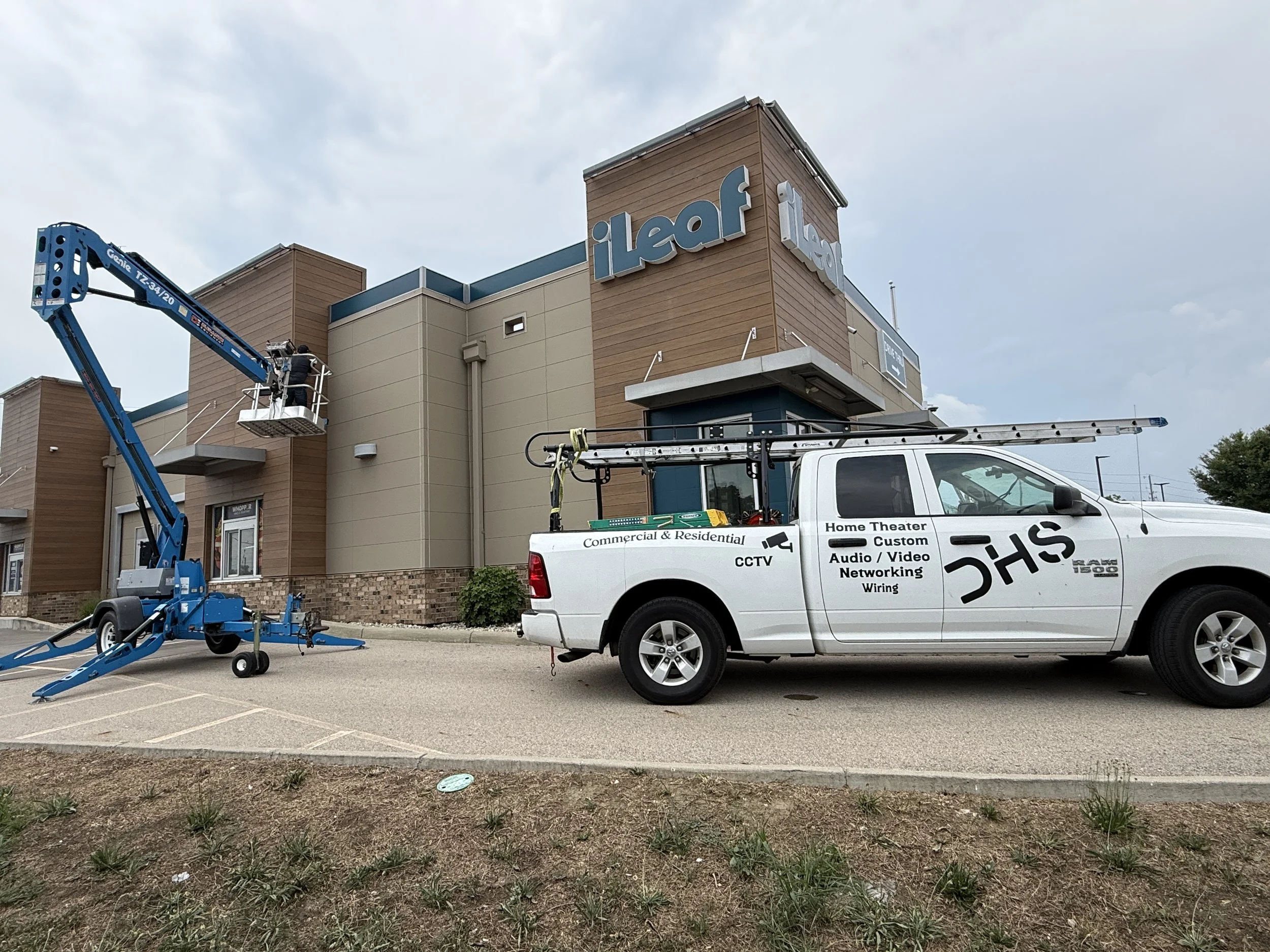 Workers using elevated lift equipment to install or maintain a logo on the exterior of a building with the sign "ilife". A white work truck parked nearby holds tools and equipment for audiovisual wiring and networking.