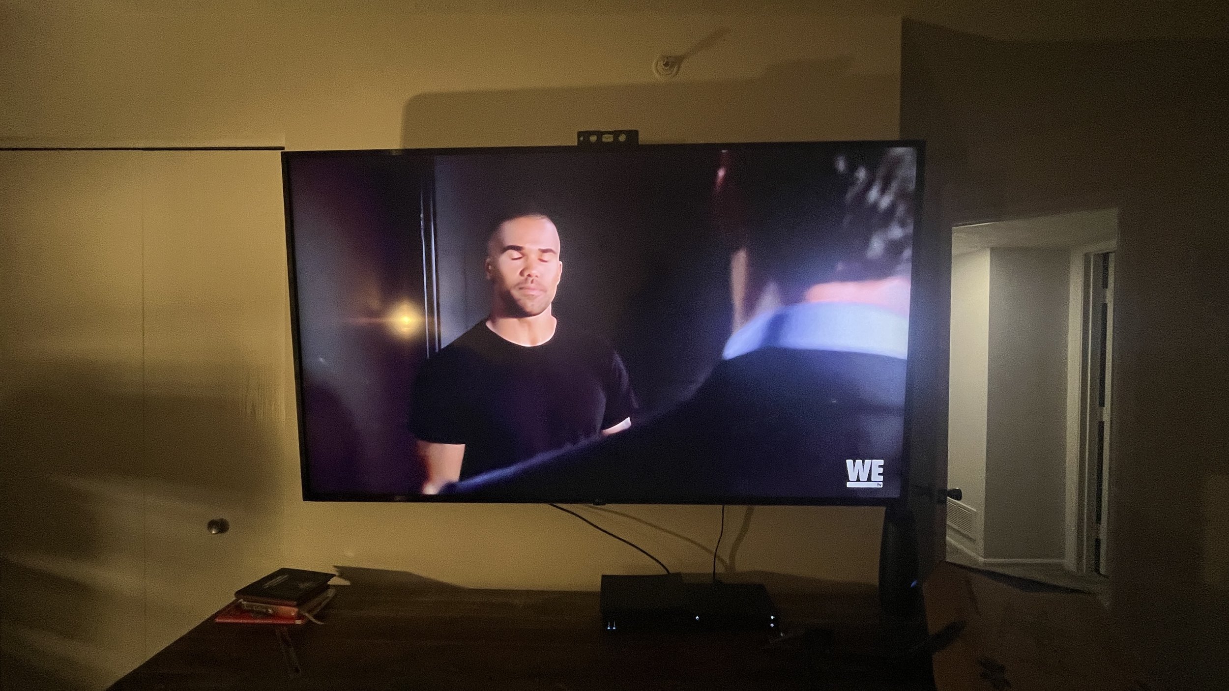 A television screen shows two men in conversation, one with eyes closed and the other turned away; the scene is from a TV show on WE Network.
