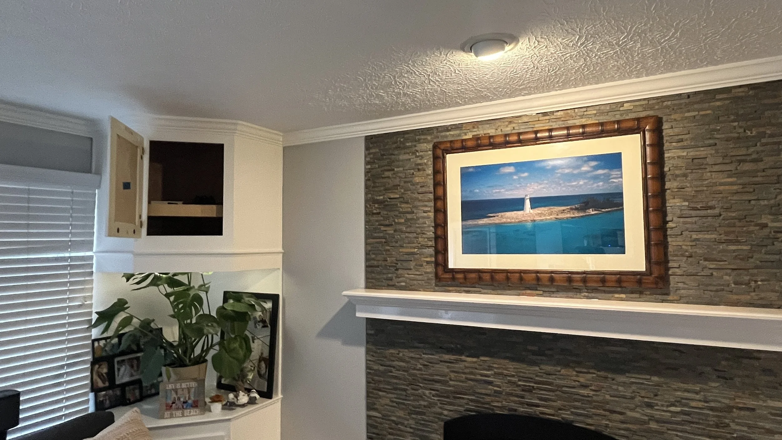 Living room interior with a framed photograph of a lighthouse on a rocky island over a stone wall, white mantel, plant with framed photos on a shelf, open cabinet with an open door, window with blinds